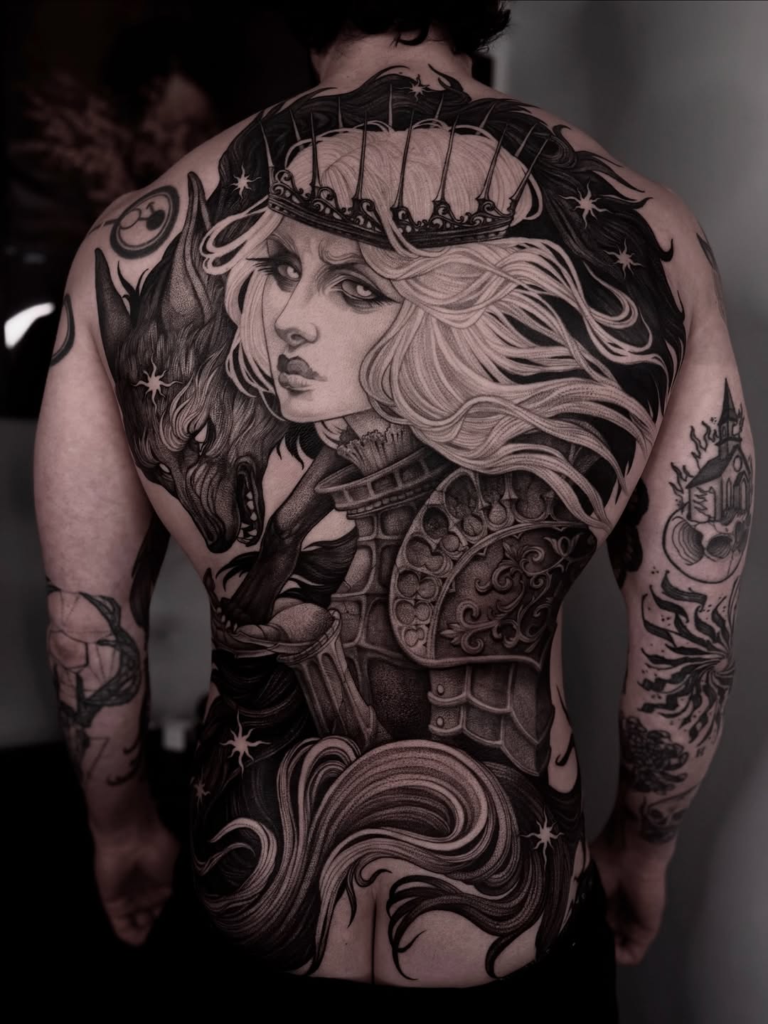 Large-scale back tattoo featuring a crowned woman with flowing hair, ornate armor, celestial elements, and decorative swirls in black ink.