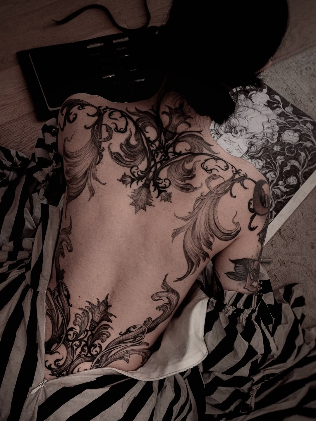Ornate scrollwork and floral tattoo covering the chest and shoulders in black ink on pale skin, displayed on striped fabric.
