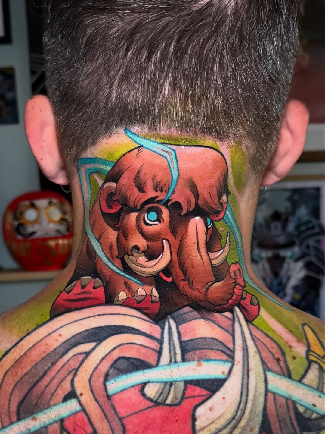 Red demon head with blue outlines and green background tattooed on the back of a neck, featuring detailed facial features and ornamental elements.