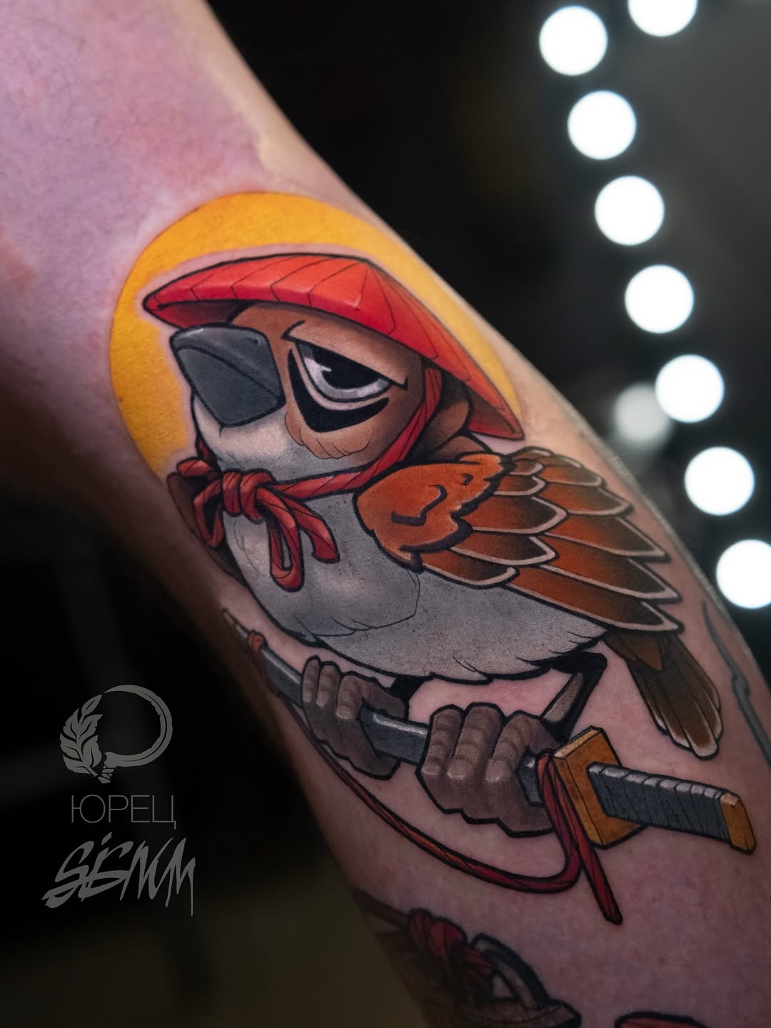 Colorful arm tattoo of a pirate bird character wearing a red hat and eye patch, holding a rifle, with a golden halo background.
