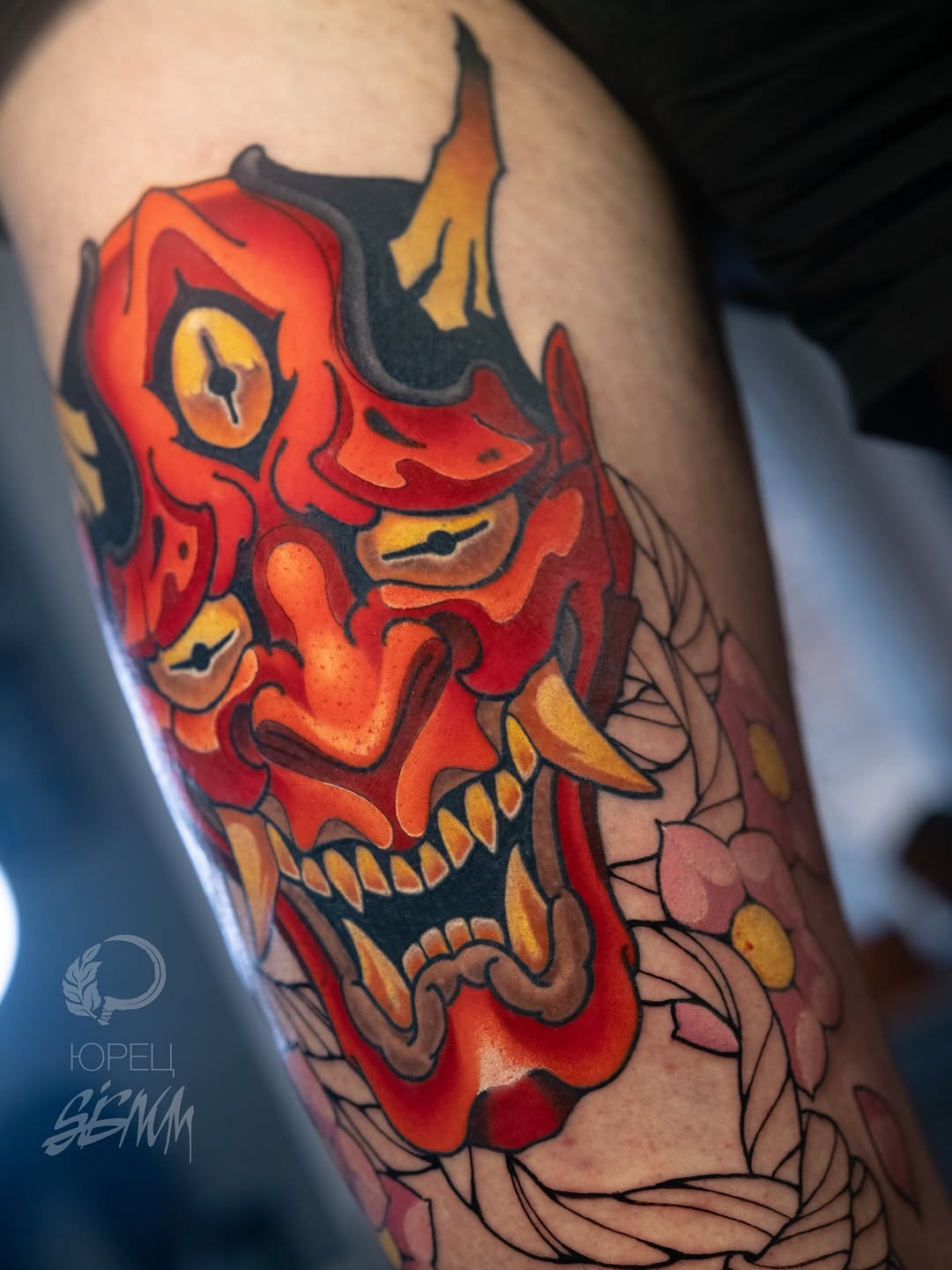 Red and gold demon mask with horns and bared teeth on upper arm, surrounded by decorative line work and floral elements.