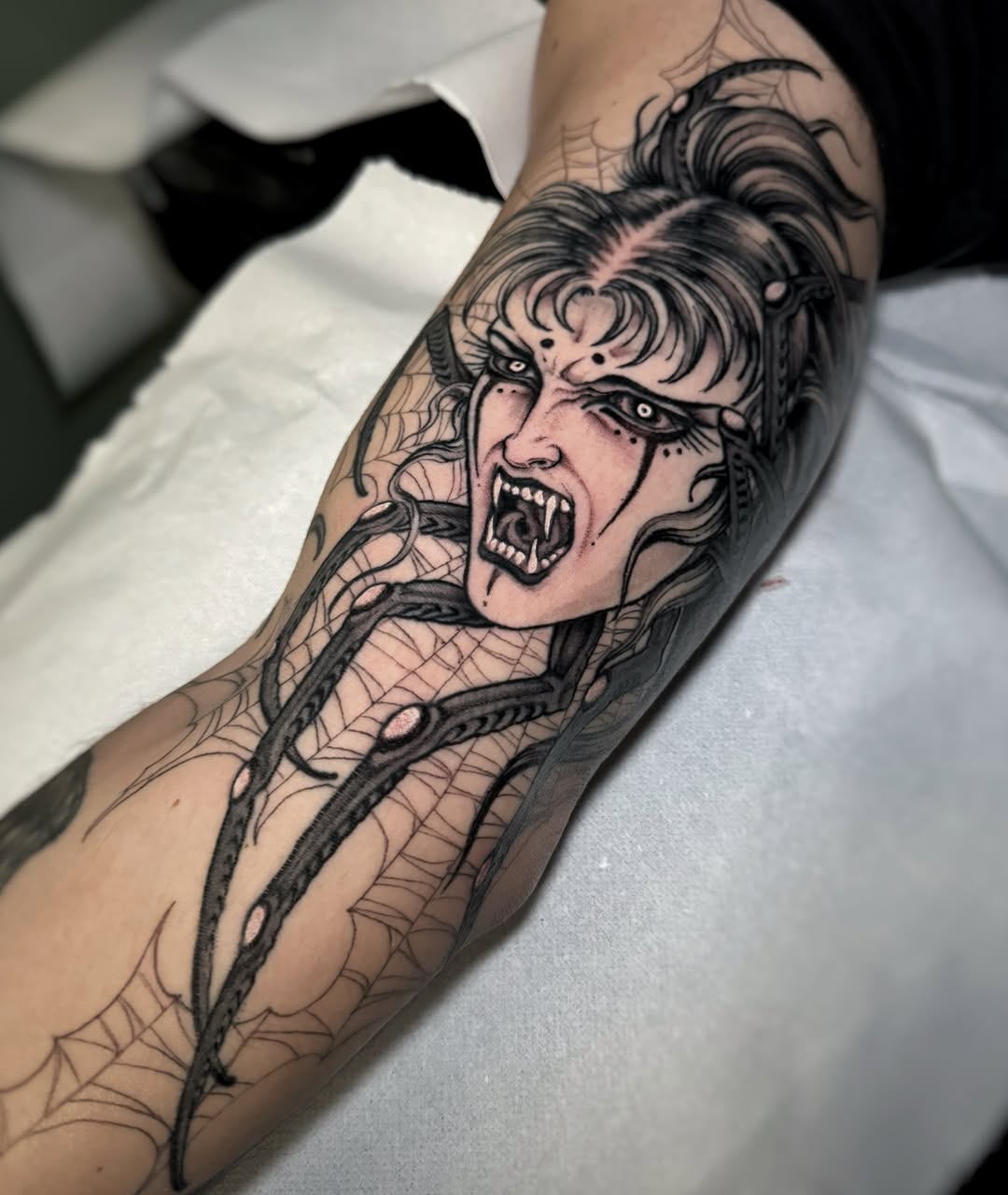 Vampire woman with fangs and horns on forearm, surrounded by web and vine details in black ink on skin.