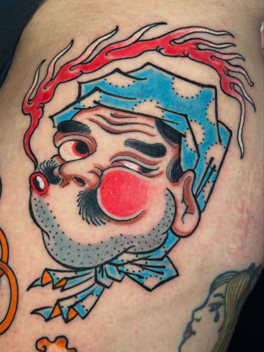 Colorful clown head tattoo on arm featuring red nose, blue hat, red flames, and gray beard with black outlines and detailed facial features.