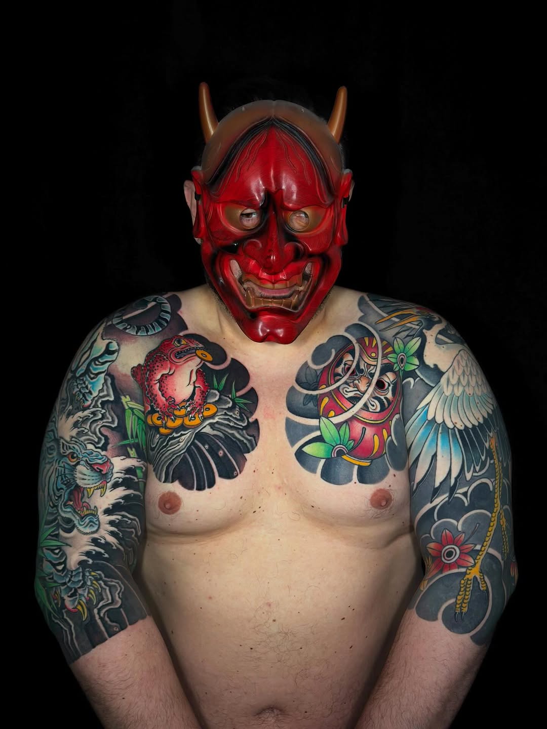Shirtless person wearing full-color sleeve tattoos featuring demons, birds, and floral motifs in red, black, and green, paired with a red horned devil mask.