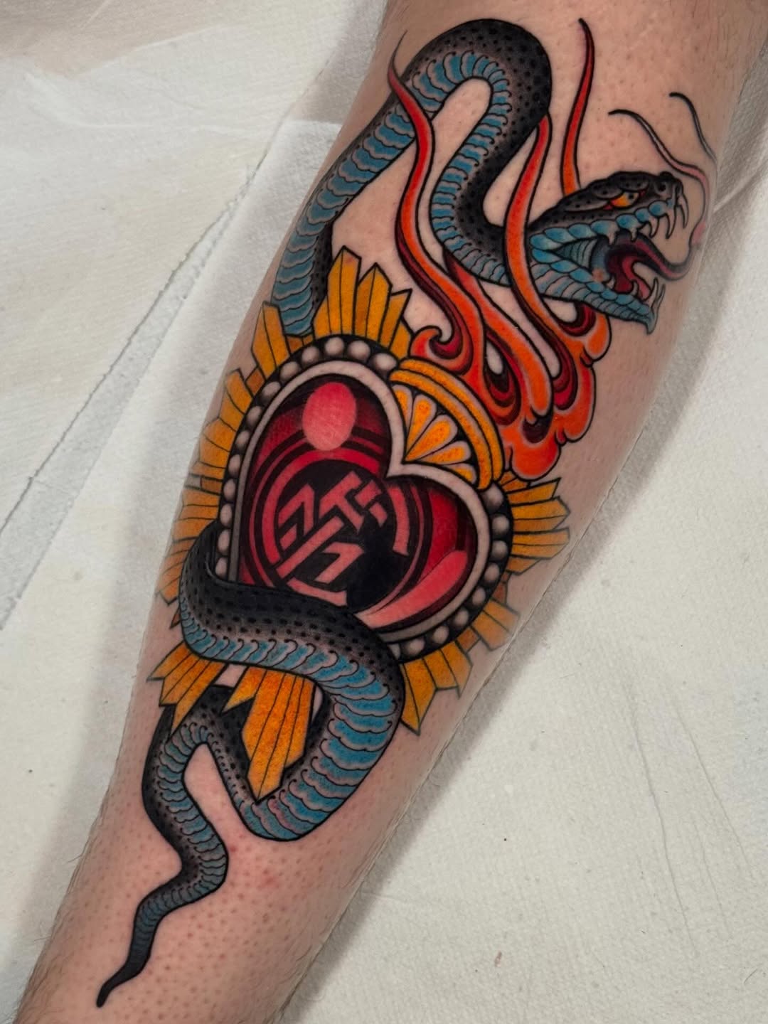 Colorful tattoo on a calf featuring two snakes intertwined around a red heart with an orange sunburst design and flames.