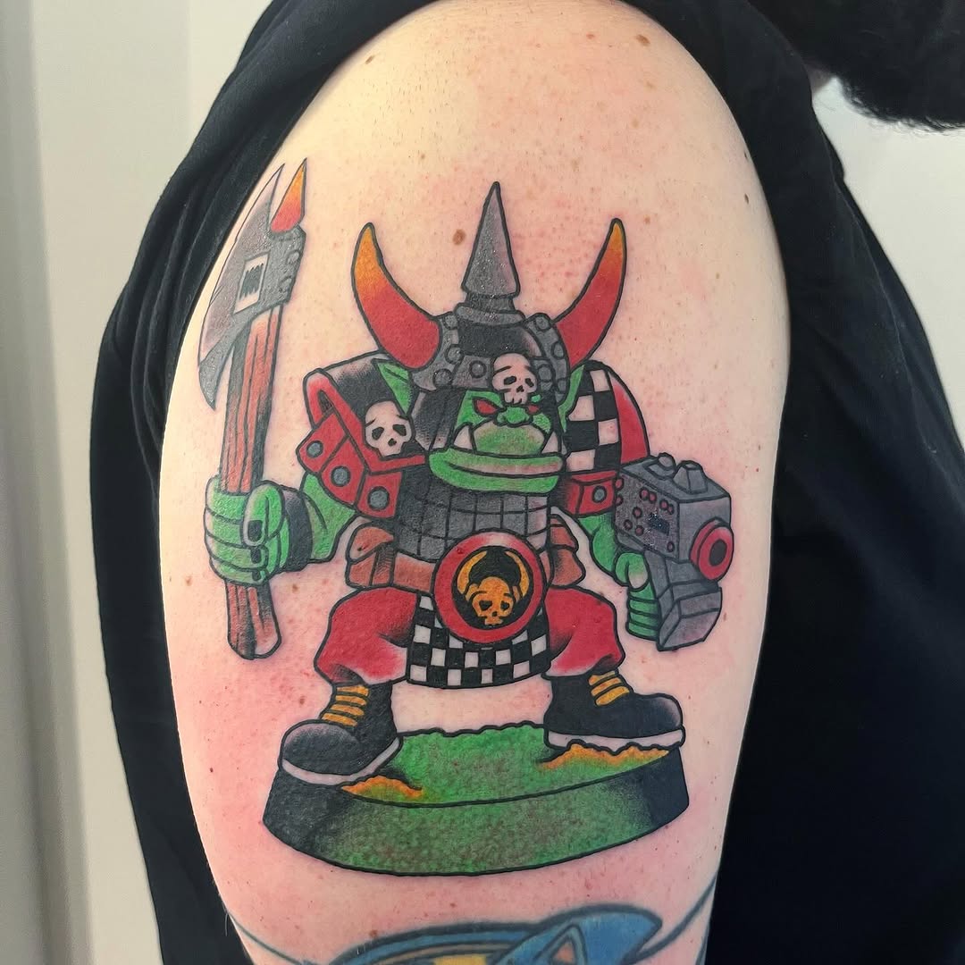 Colorful demon warrior tattoo on upper arm featuring red horns, green skin, black armor, skull chest emblem, weapons, and green base platform.