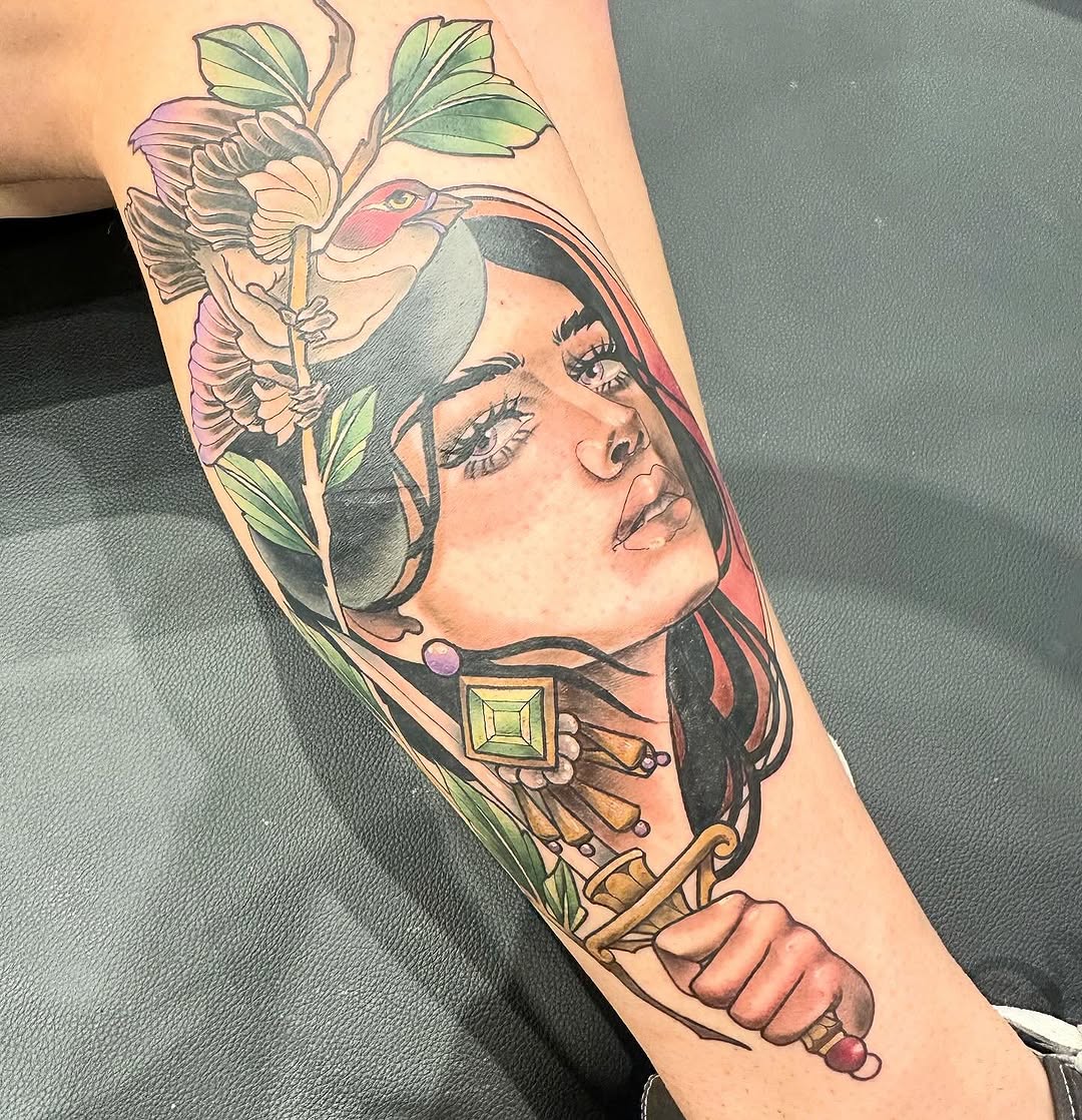 Colorful forearm tattoo featuring a woman's face with closed eyes, a bird perched on flowers, decorative gems, and ornamental elements in pink, green, and gold tones.