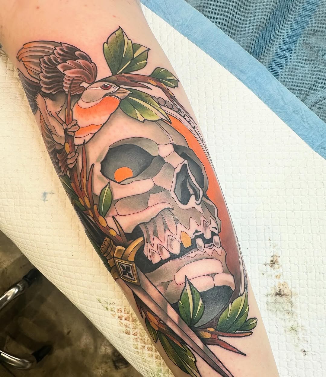 Colorful tattoo on forearm featuring a skull with bird, green foliage, and orange accents arranged vertically on the arm.