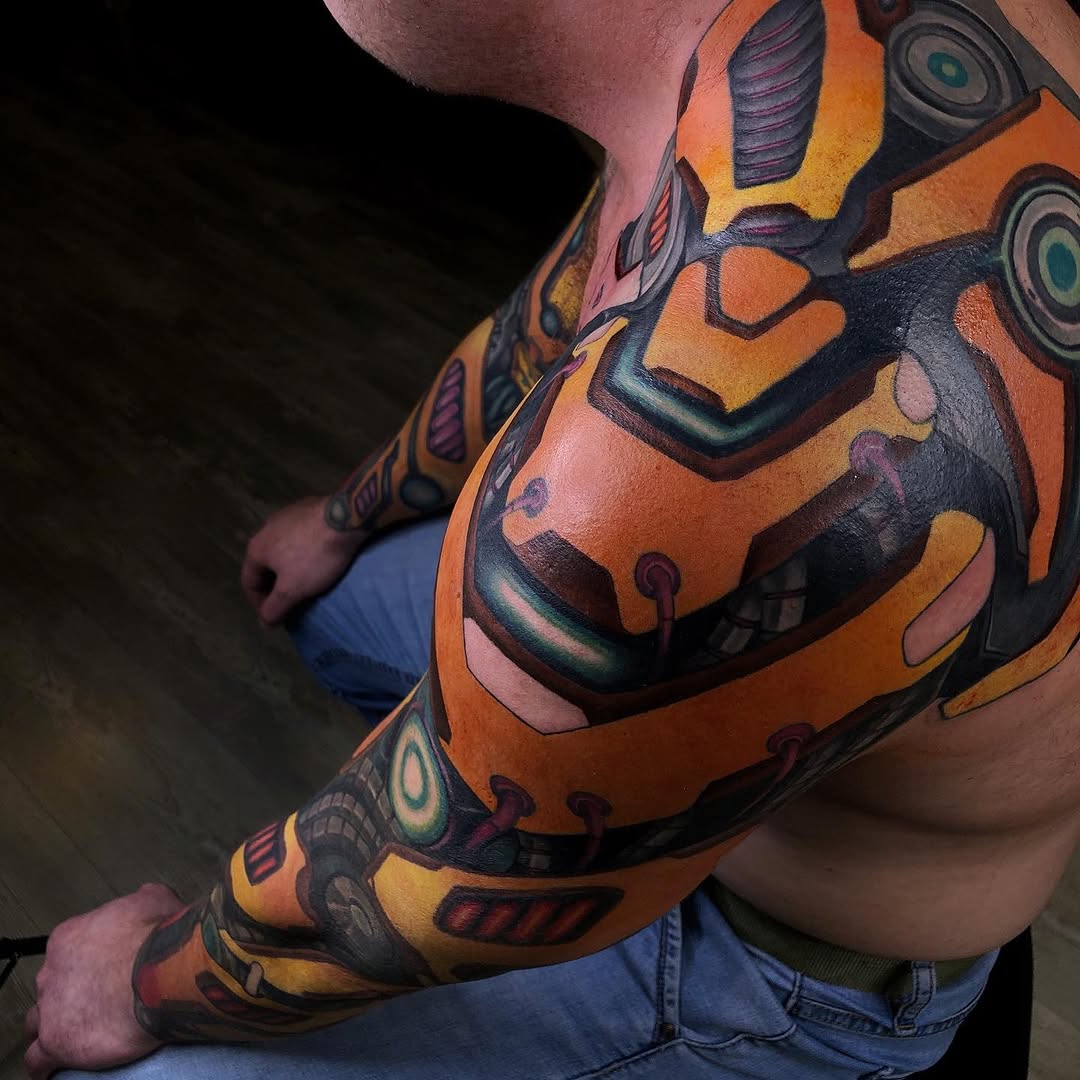 Colorful full-sleeve tattoo featuring orange, black, and yellow geometric and mechanical designs with circular eye-like elements on a person's arm.