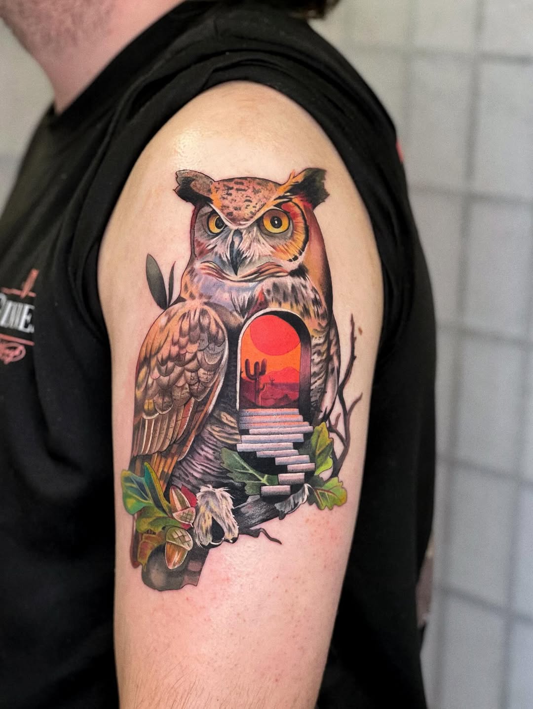 Colored owl tattoo on upper arm with golden eyes, desert sunset doorway in chest, surrounding branches and green foliage details.