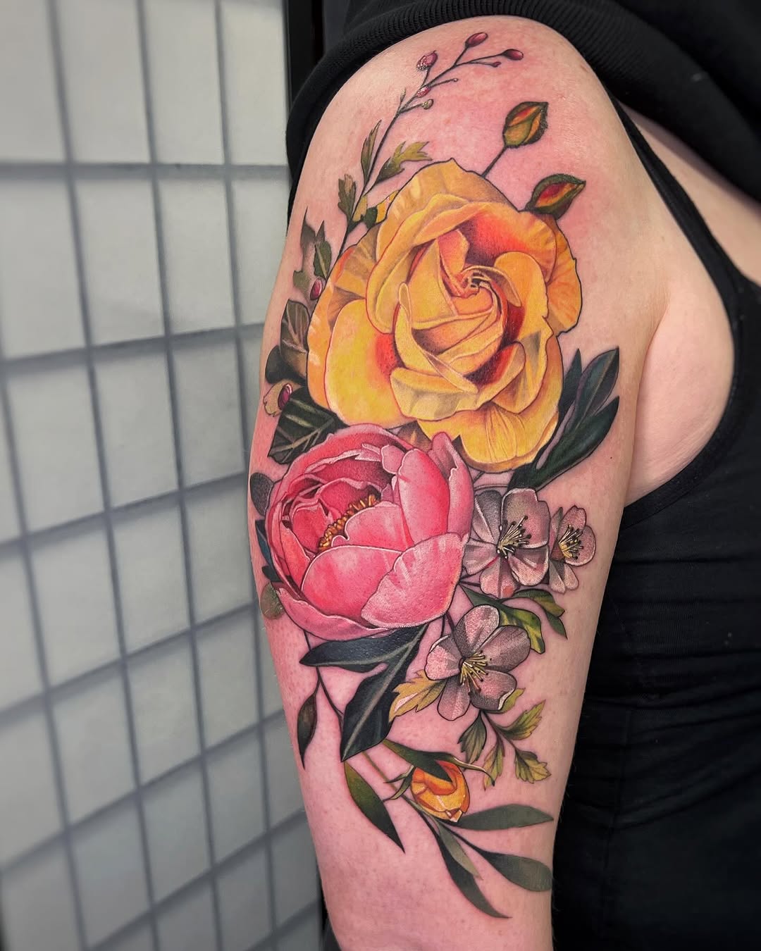 Colorful floral tattoo on upper arm featuring a yellow rose, pink peony, and small purple flowers with green leaves and buds on pink skin.