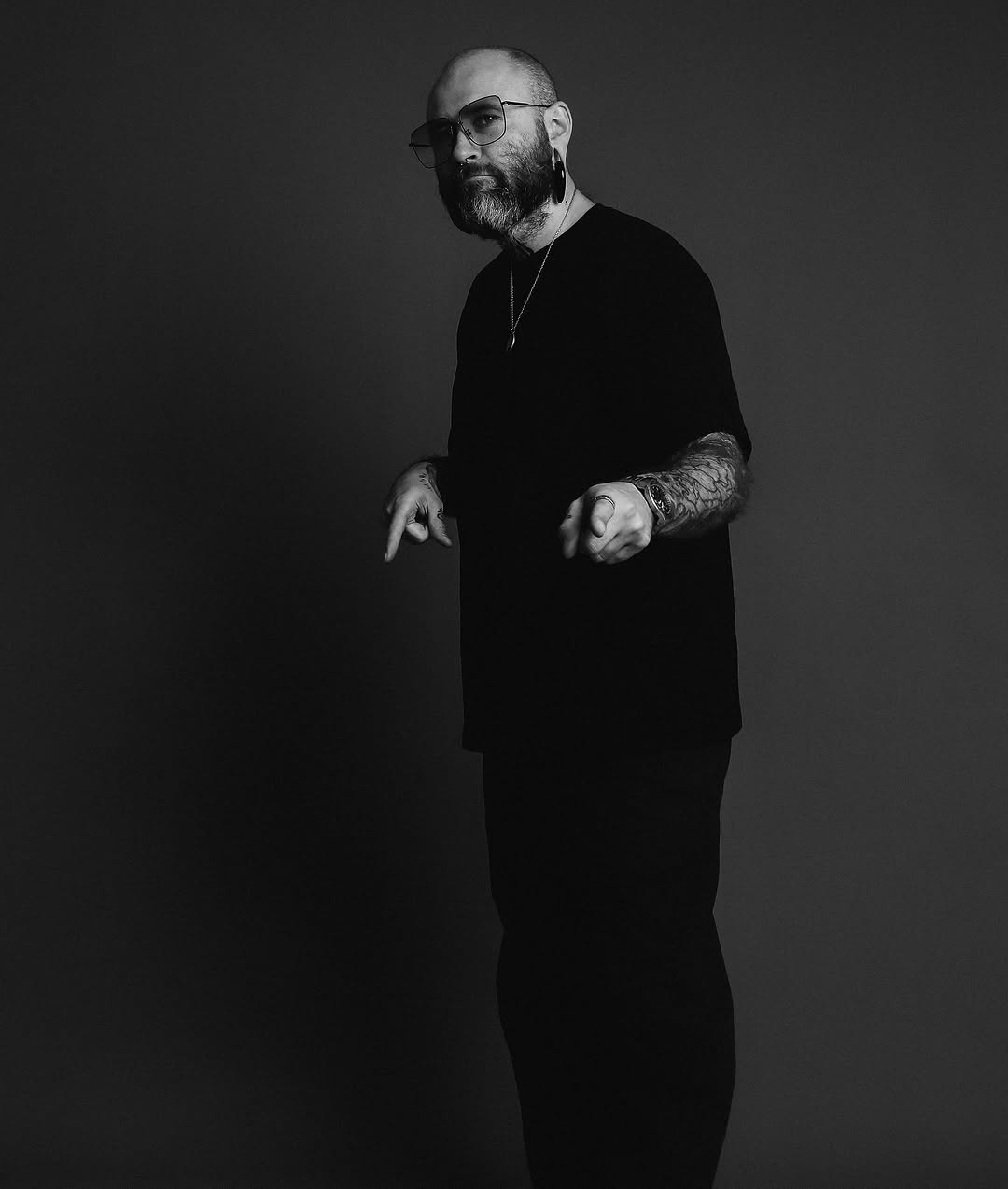 Bald man with full beard wearing glasses and black long-sleeved shirt displays large detailed tattoo covering his entire right forearm and hand against dark background.