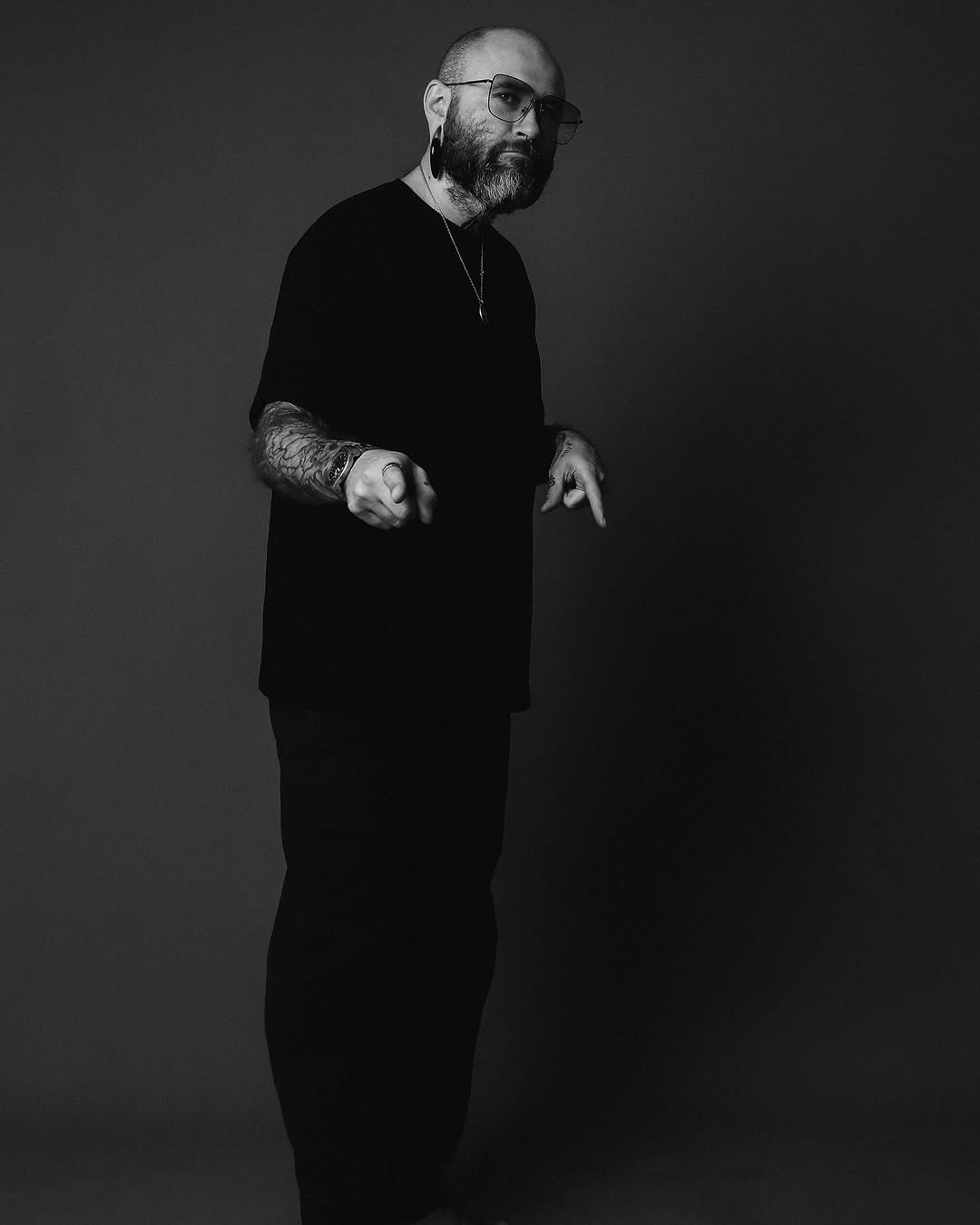Black and white studio portrait of a bearded man wearing glasses and dark clothing, displaying an arm tattoo on his left sleeve.