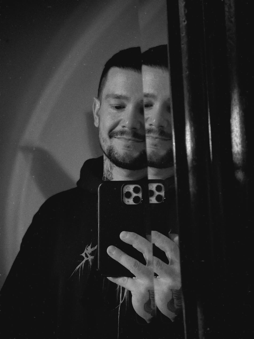 Man with a spiky star tattoo on his chest takes a mirror selfie in black and white, holding a smartphone.