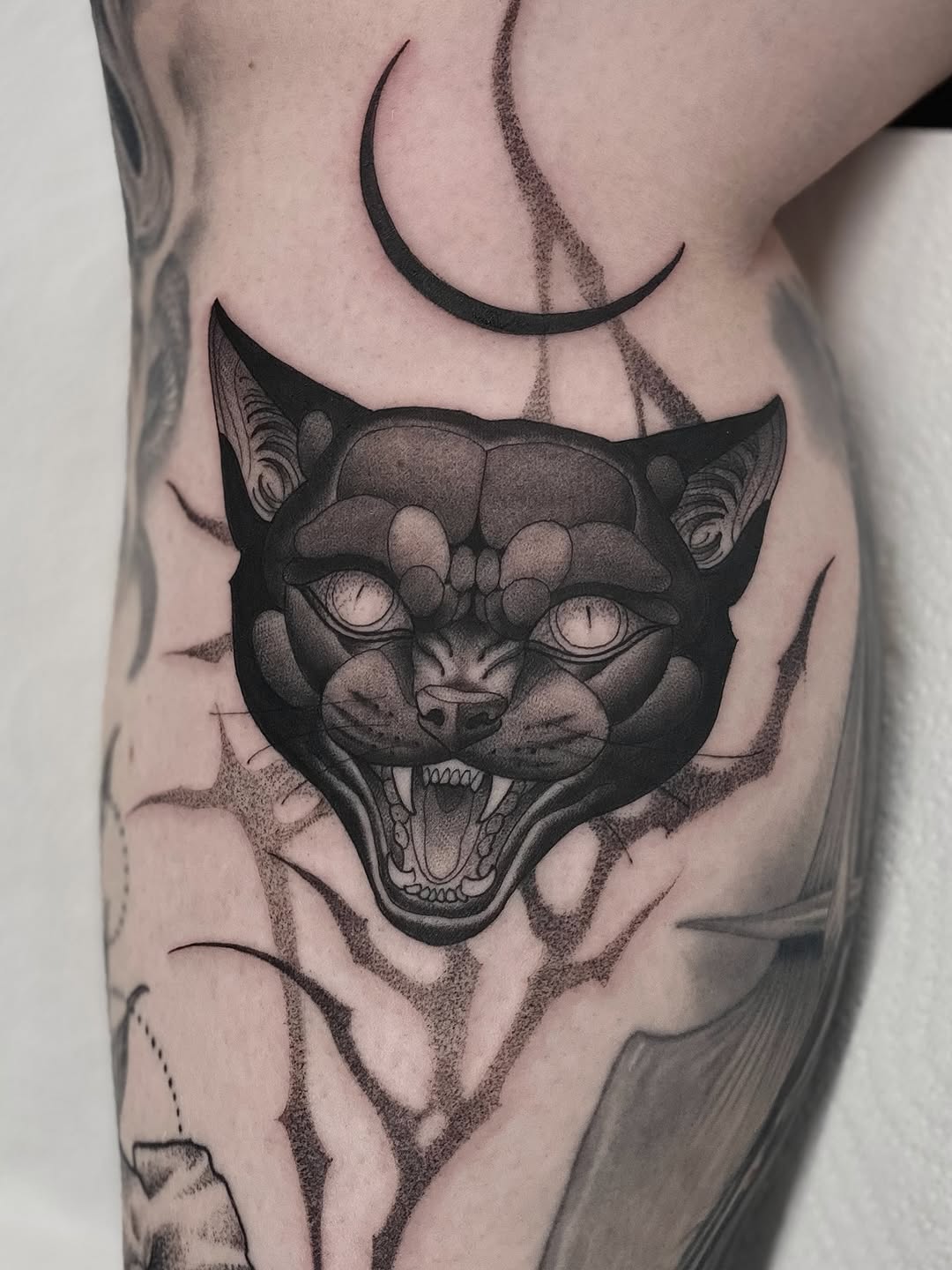 Demonic cat head with bared fangs and crescent moons tattooed on upper leg in black ink with shading and dotwork details.