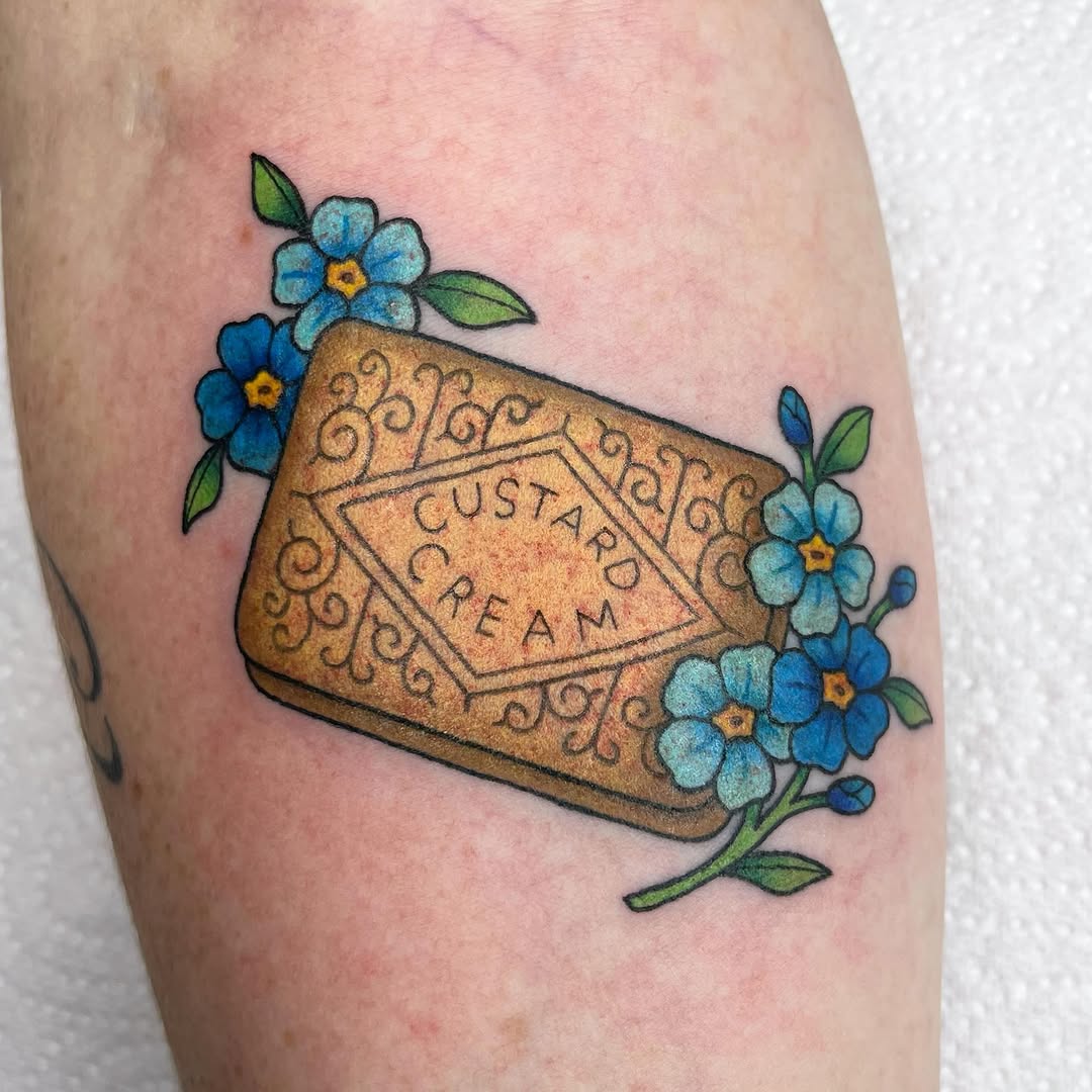 Custard Cream biscuit package tattoo with blue forget-me-not flowers and green leaves on the upper arm.