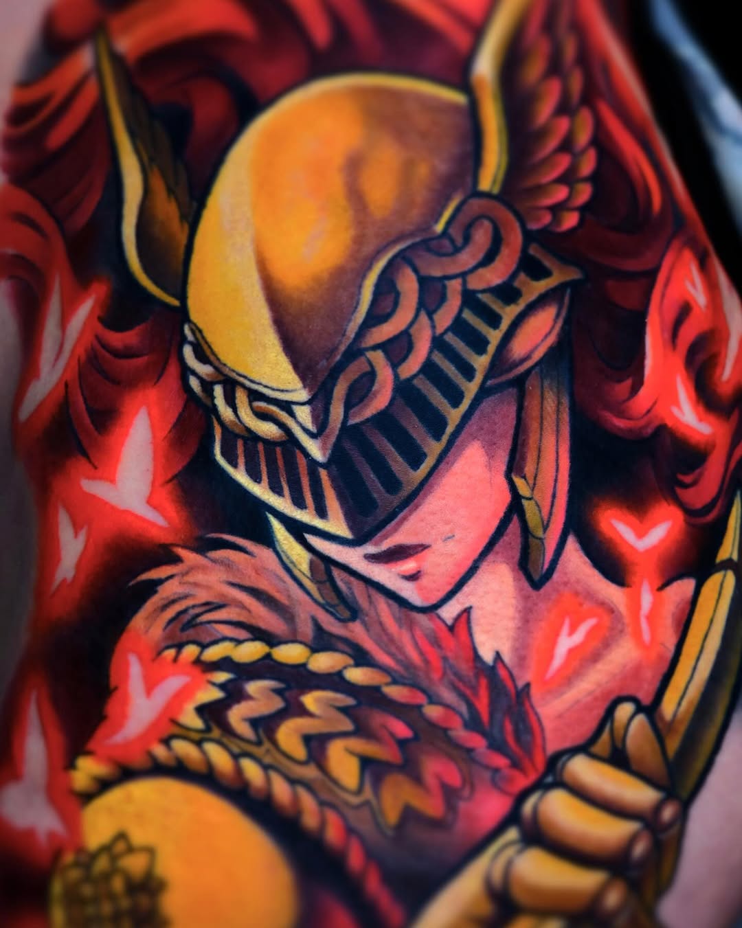 Colorful shoulder tattoo featuring a winged figure in gold, red, and orange tones with ornate armor details and flowing hair.