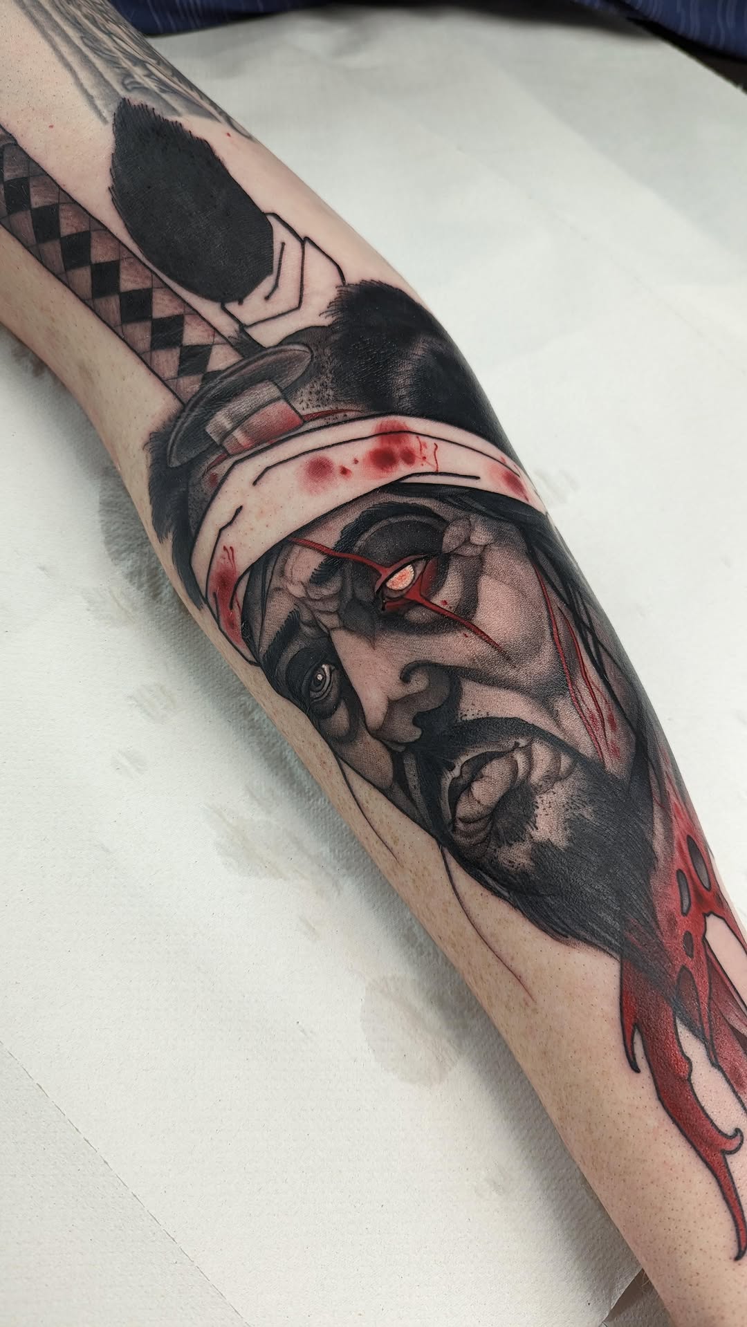 Arm tattoo featuring a bearded man's profile with bandaged head and red eyes, accompanied by geometric patterns and red dripping elements.