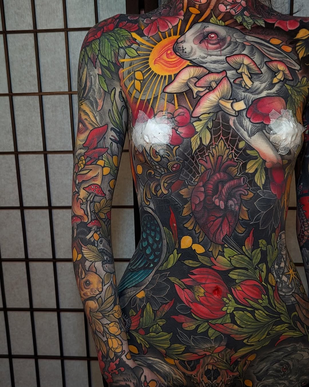 Full-body tattoo featuring a rabbit skull with mushrooms, flowers, and botanical elements in red, yellow, green, and black ink on chest and arms.