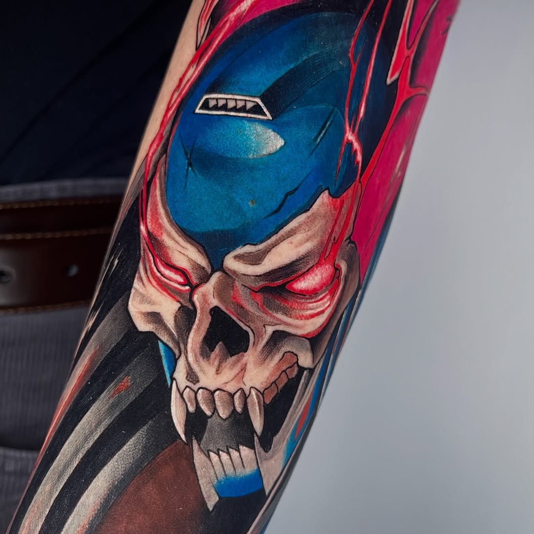 Colorful arm tattoo featuring a blue-masked superhero face merged with a skull, rendered in red, blue, black, and flesh tones.