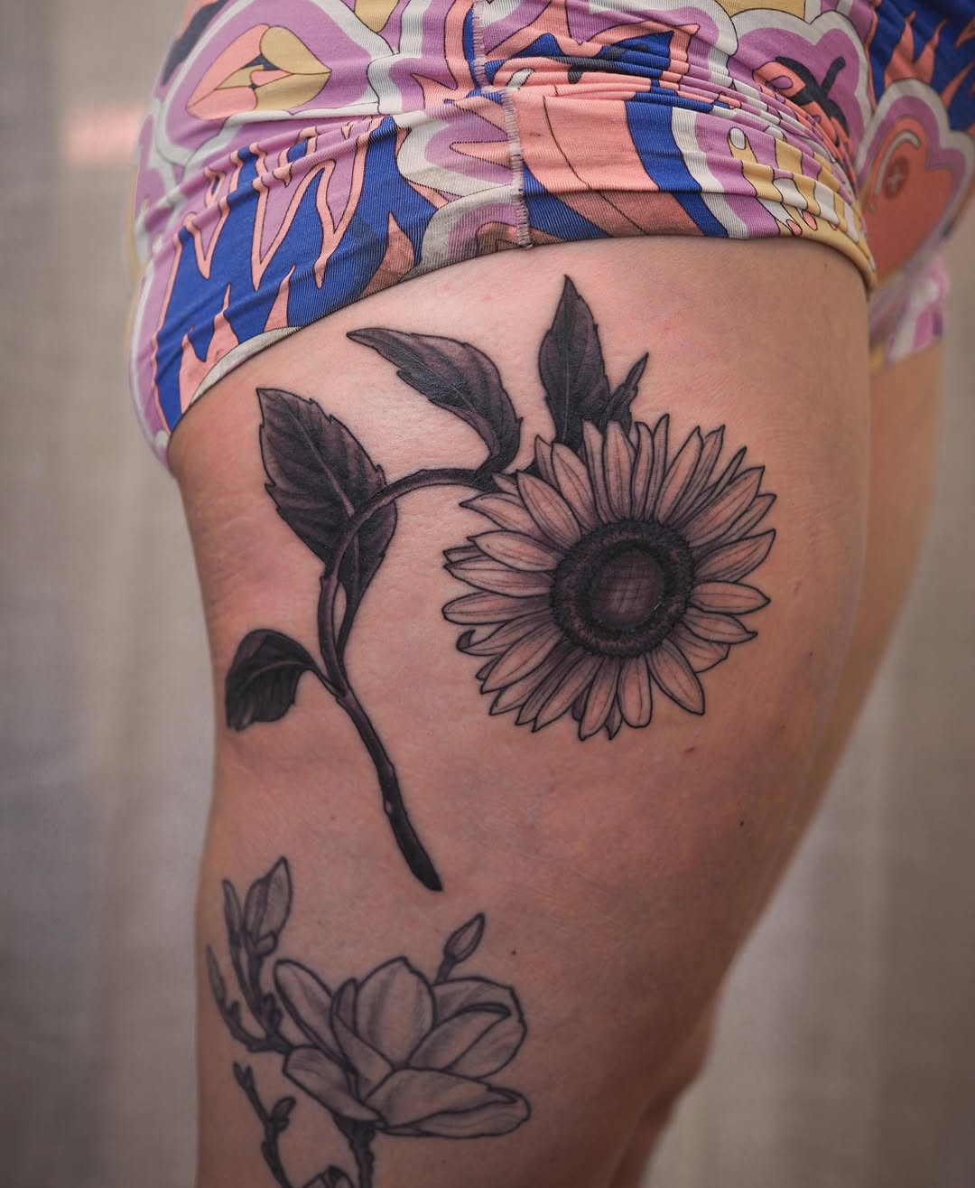 Sunflower with stem and leaves tattooed on thigh in black ink with pink shading, paired with a lower floral design.
