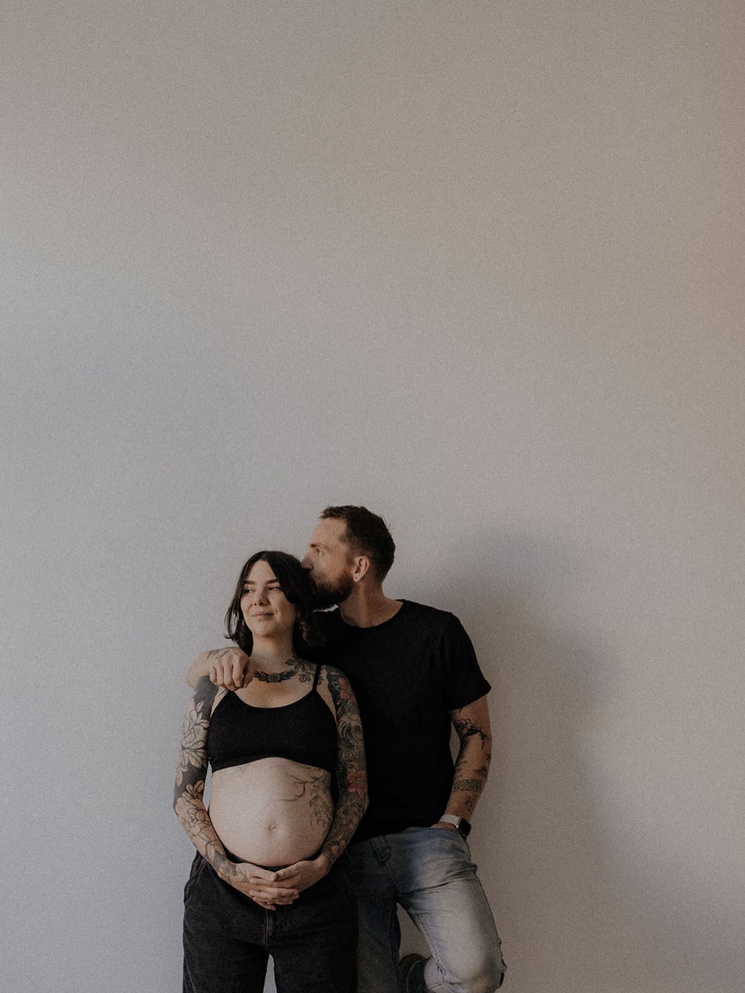Tattooed couple posing against a white wall, woman with extensive arm tattoos cradling her pregnant belly, man standing behind with arm tattoos visible.