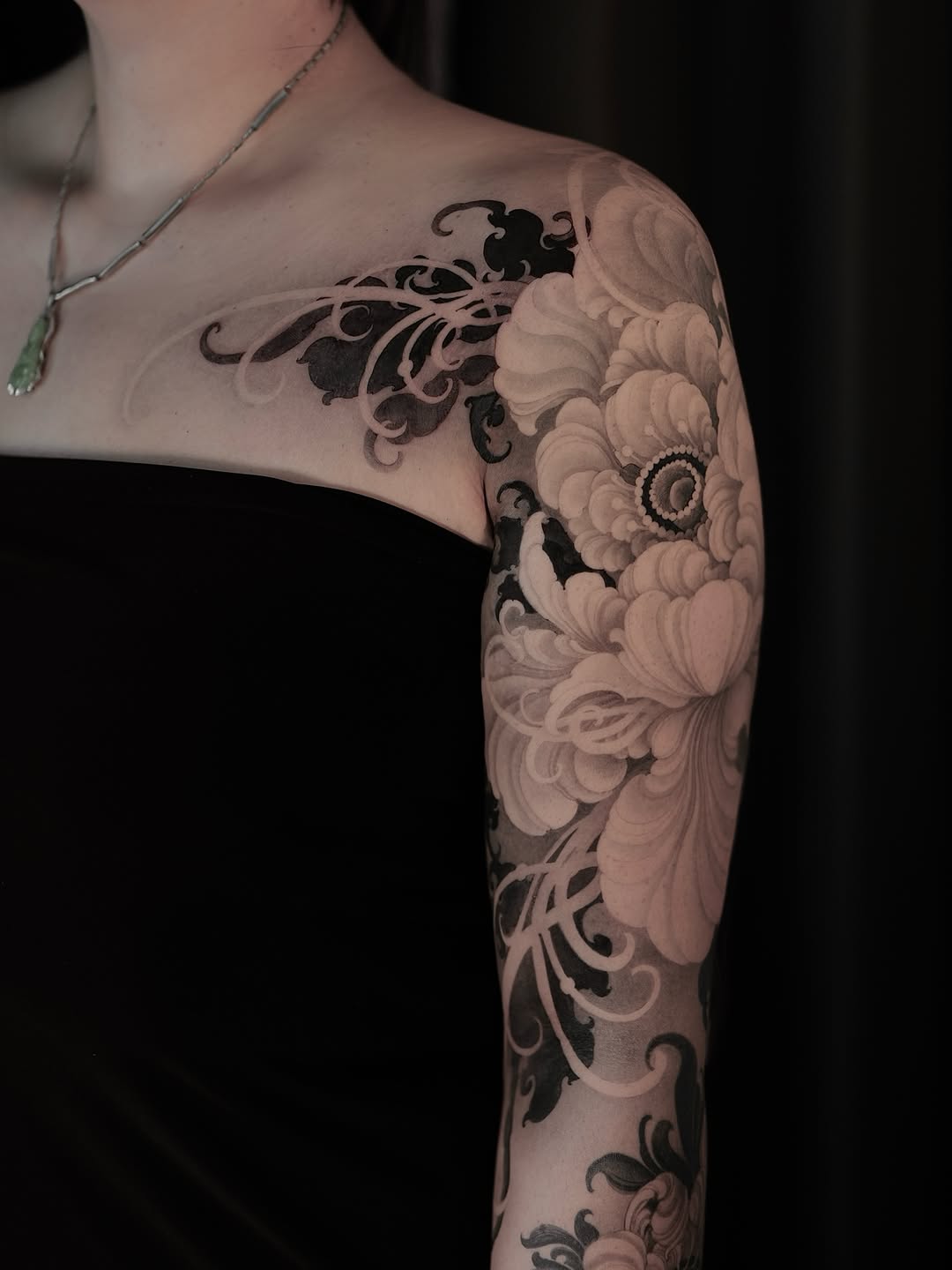 Full arm sleeve tattoo featuring ornate scrollwork and large peony flowers in black and gray ink on pale skin.