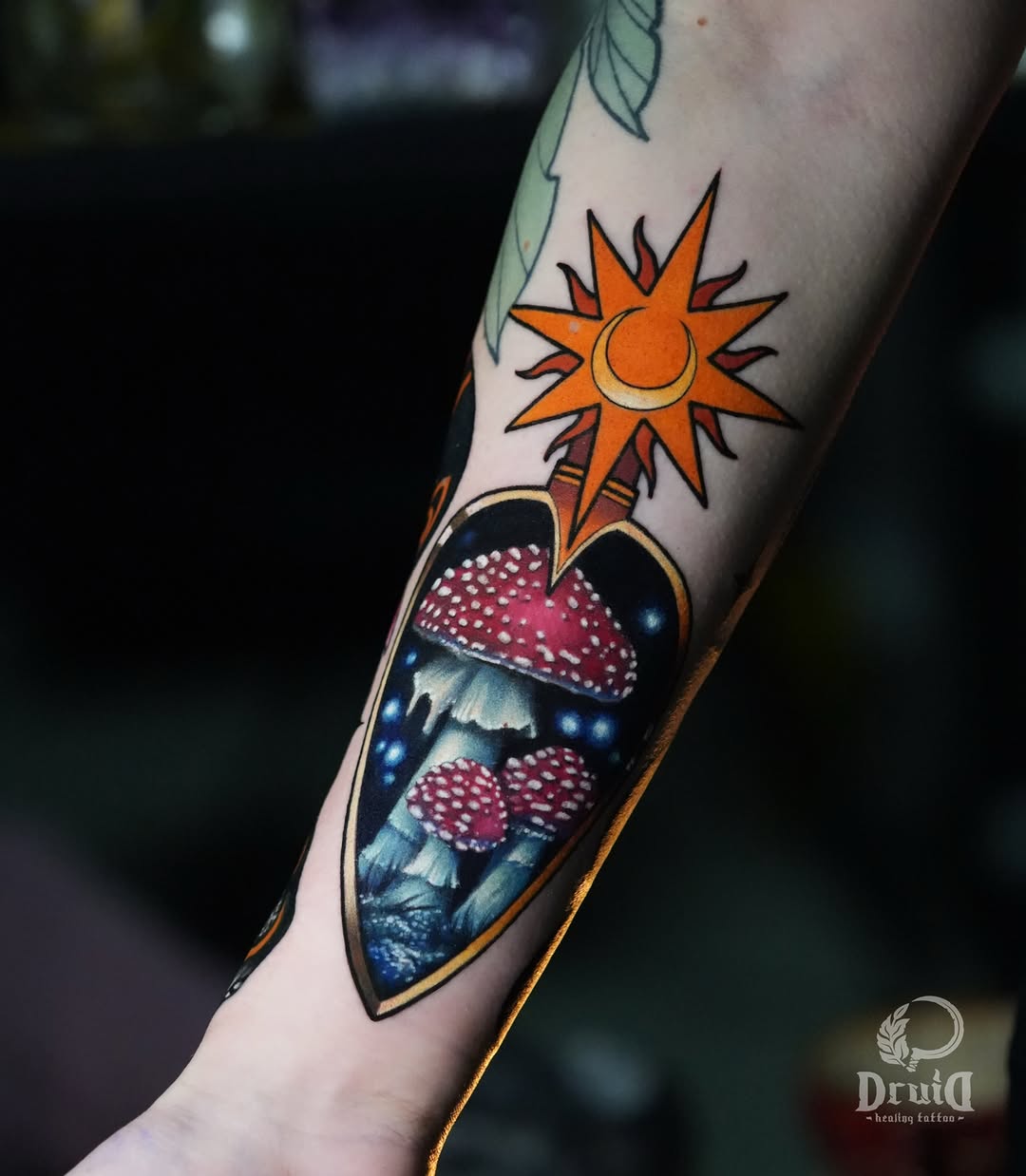 Colorful arm tattoo featuring an orange sun with crescent moon above a teardrop-shaped frame containing red mushrooms and blue forest landscape.