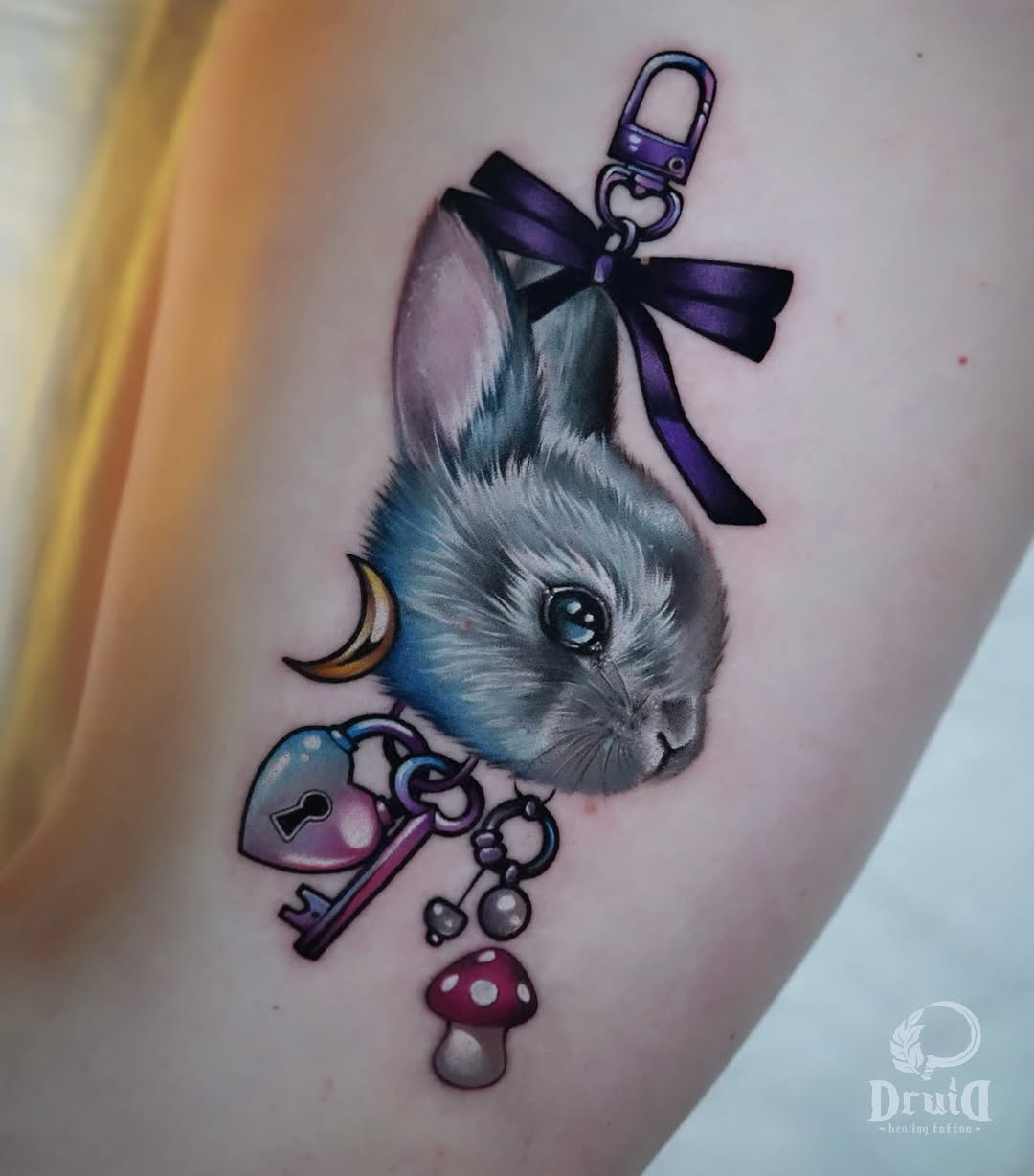 Colorful shoulder tattoo featuring a gray cat's head with purple bow and chain, accompanied by a heart-shaped lock, key, moon, and mushroom charm.