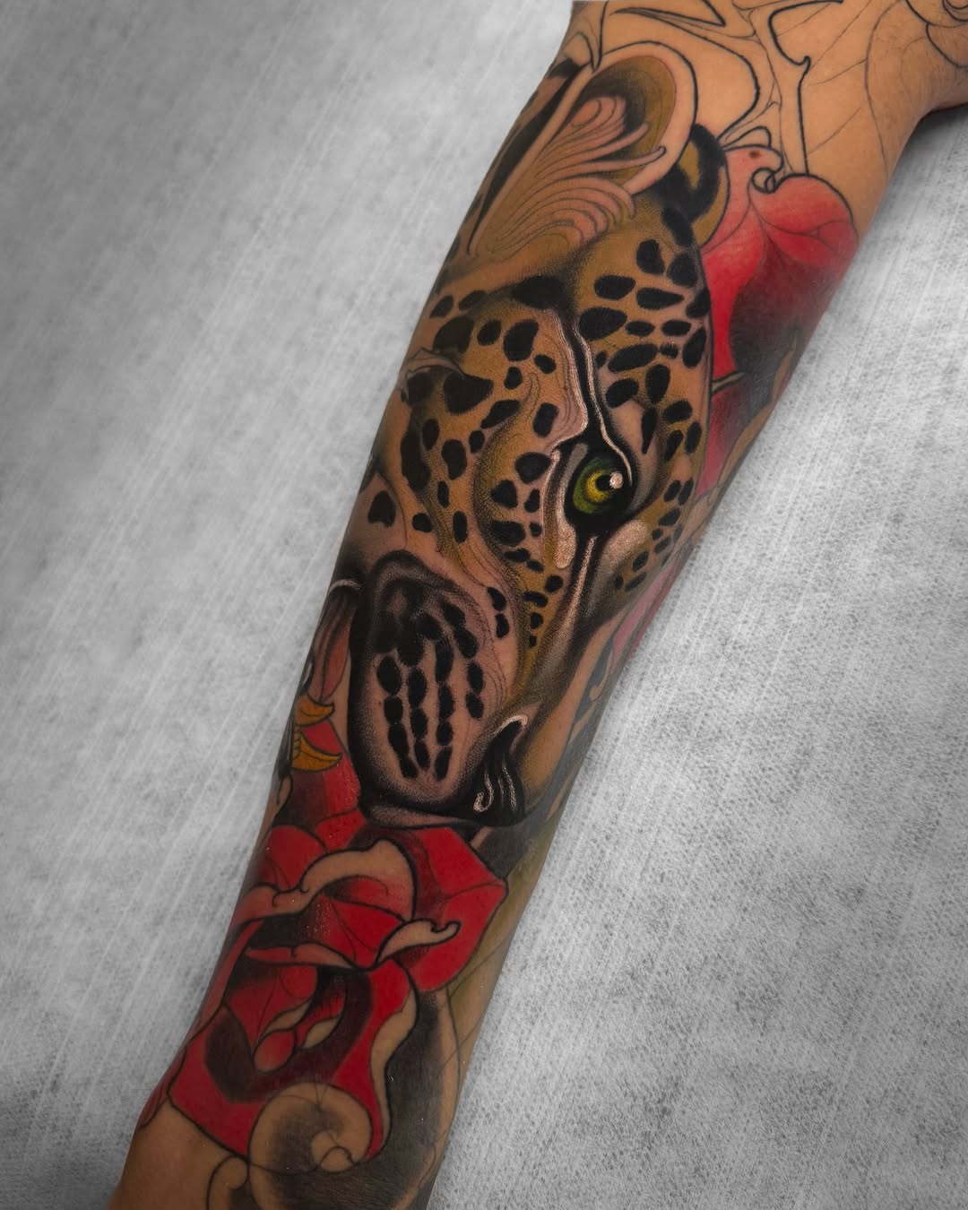 Leopard head tattoo with yellow eyes and spotted coat on forearm, surrounded by red roses and ornamental designs in brown and red tones.