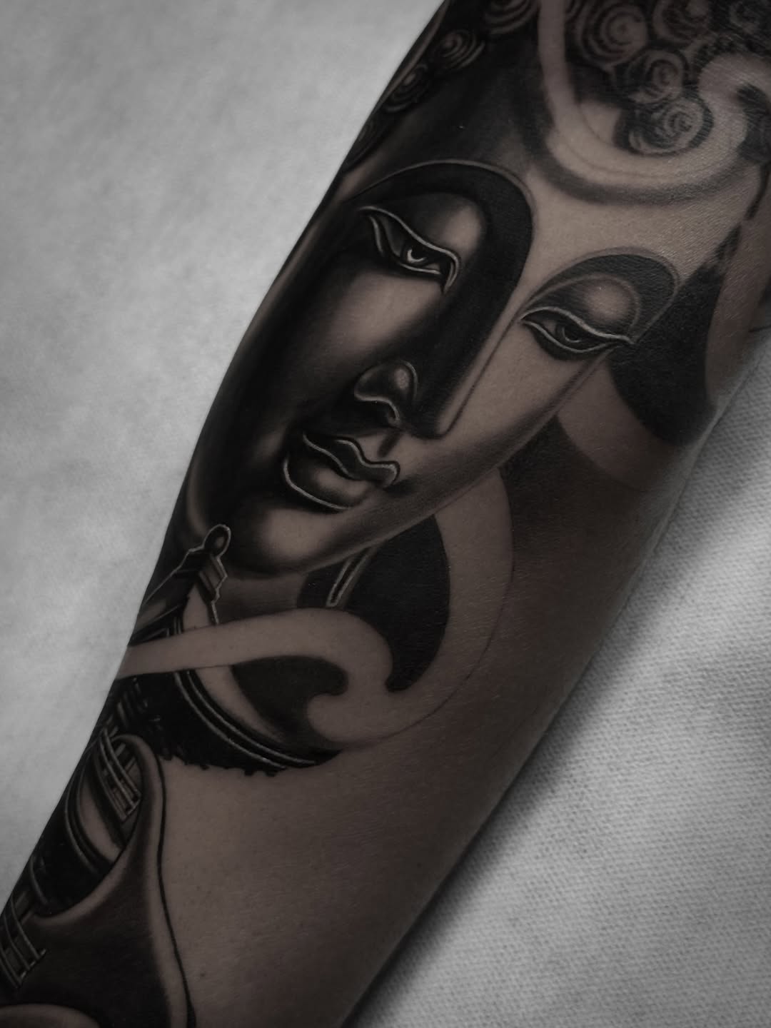 Buddha faces with closed eyes and ornamental headdress details tattooed in black ink on an upper arm, shown against white fabric.