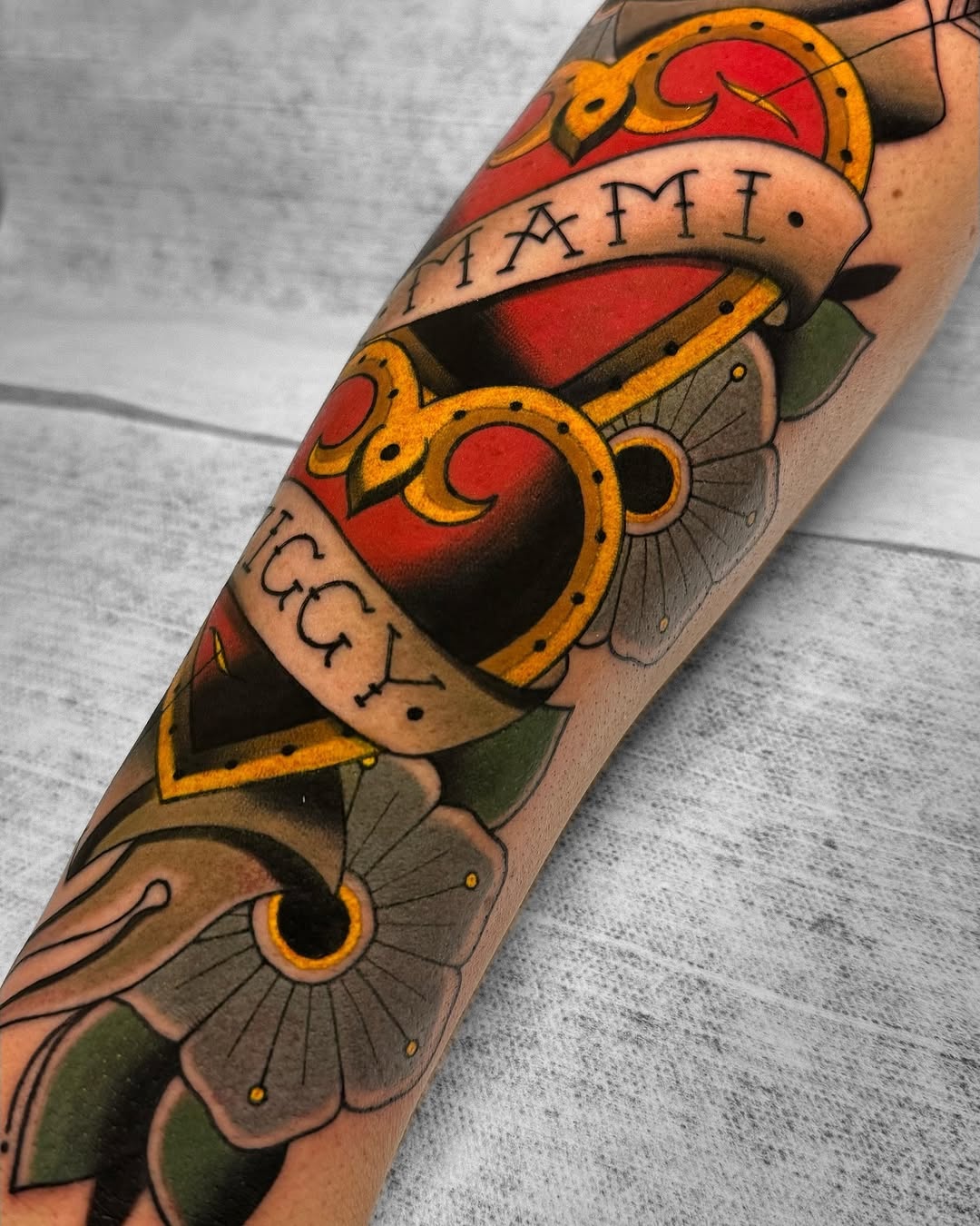Colorful tattoo on forearm featuring a red heart with gold ornamental frame, banner with text, and green floral elements below.