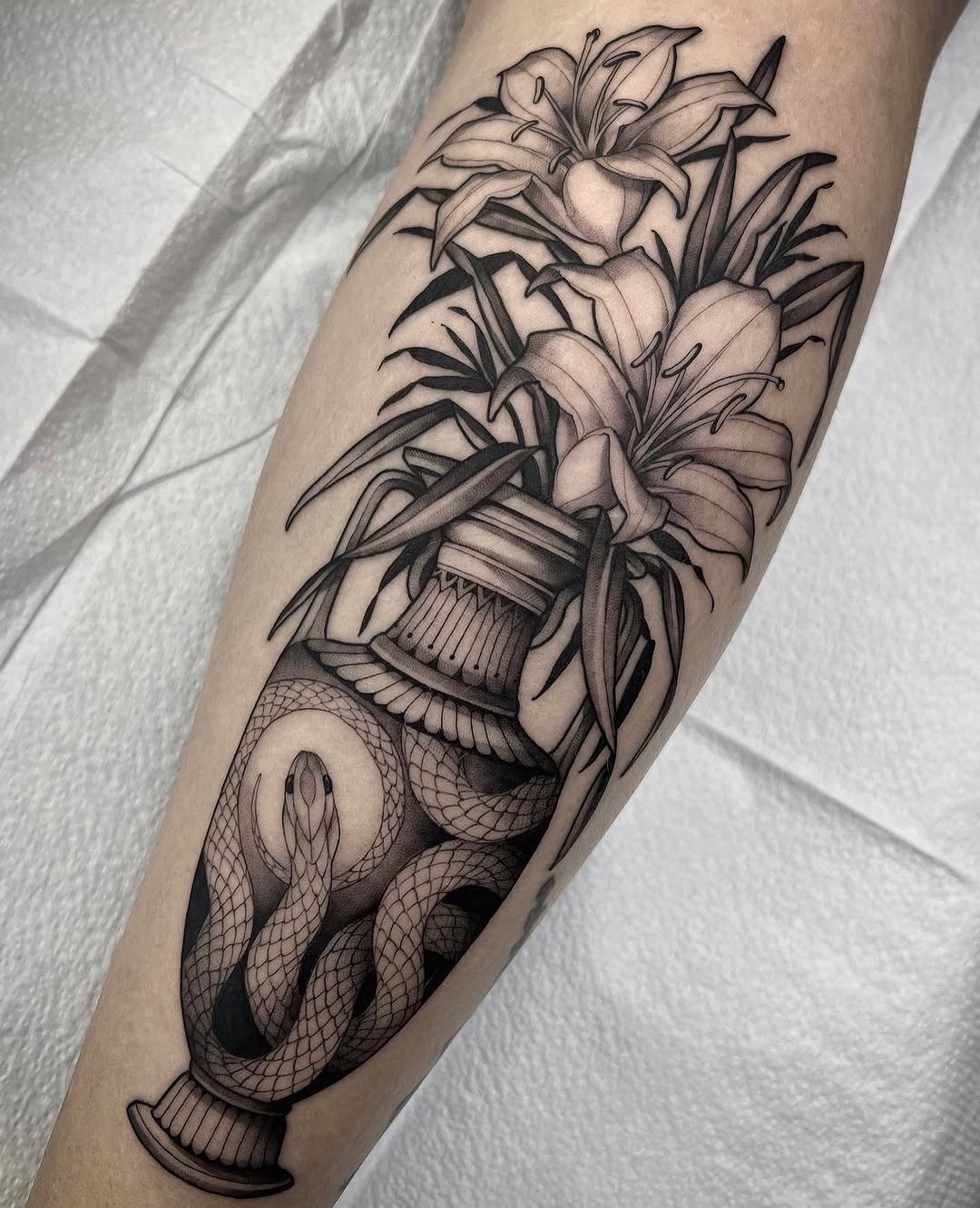 Forearm tattoo featuring lilies in a vase with a coiled snake, rendered in black ink with shading on light skin.