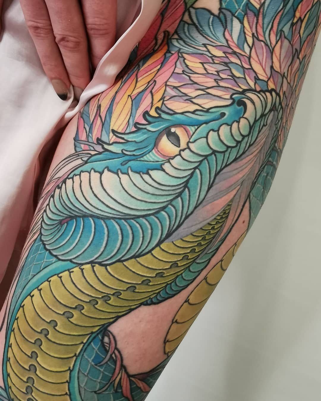 Colorful dragon wing tattoo on an arm featuring turquoise, pink, yellow, and gold scales with detailed feather work and black outlines.