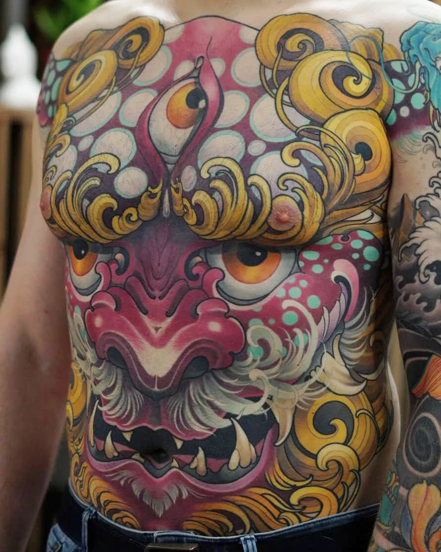 Colorful chest tattoo featuring a stylized mask with red and pink tones, golden ornamental scrollwork, large expressive eyes, and decorative circular patterns.