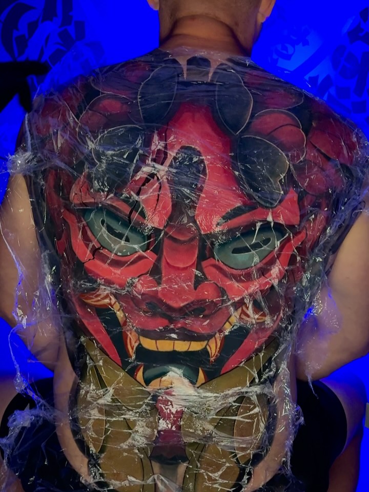 Back tattoo featuring a large red demonic face with green eyes and yellow teeth, surrounded by black geometric line work and additional facial designs.