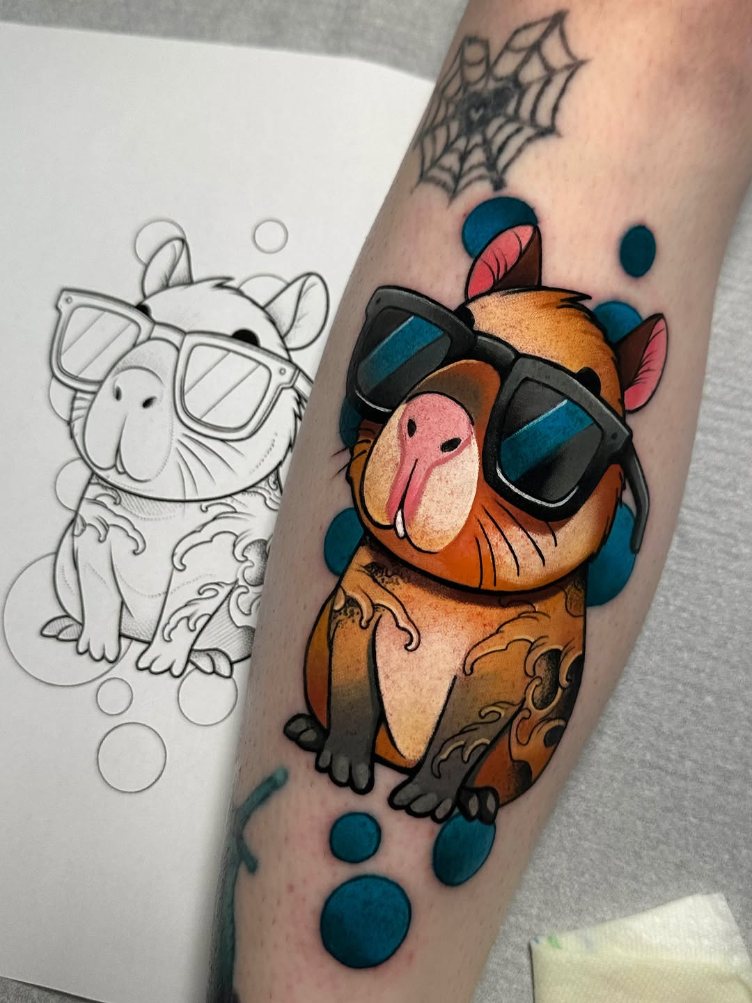 Lucky cat tattoo on forearm featuring brown and orange coloring with black sunglasses, blue accents, decorative circles, and a spider web detail above.