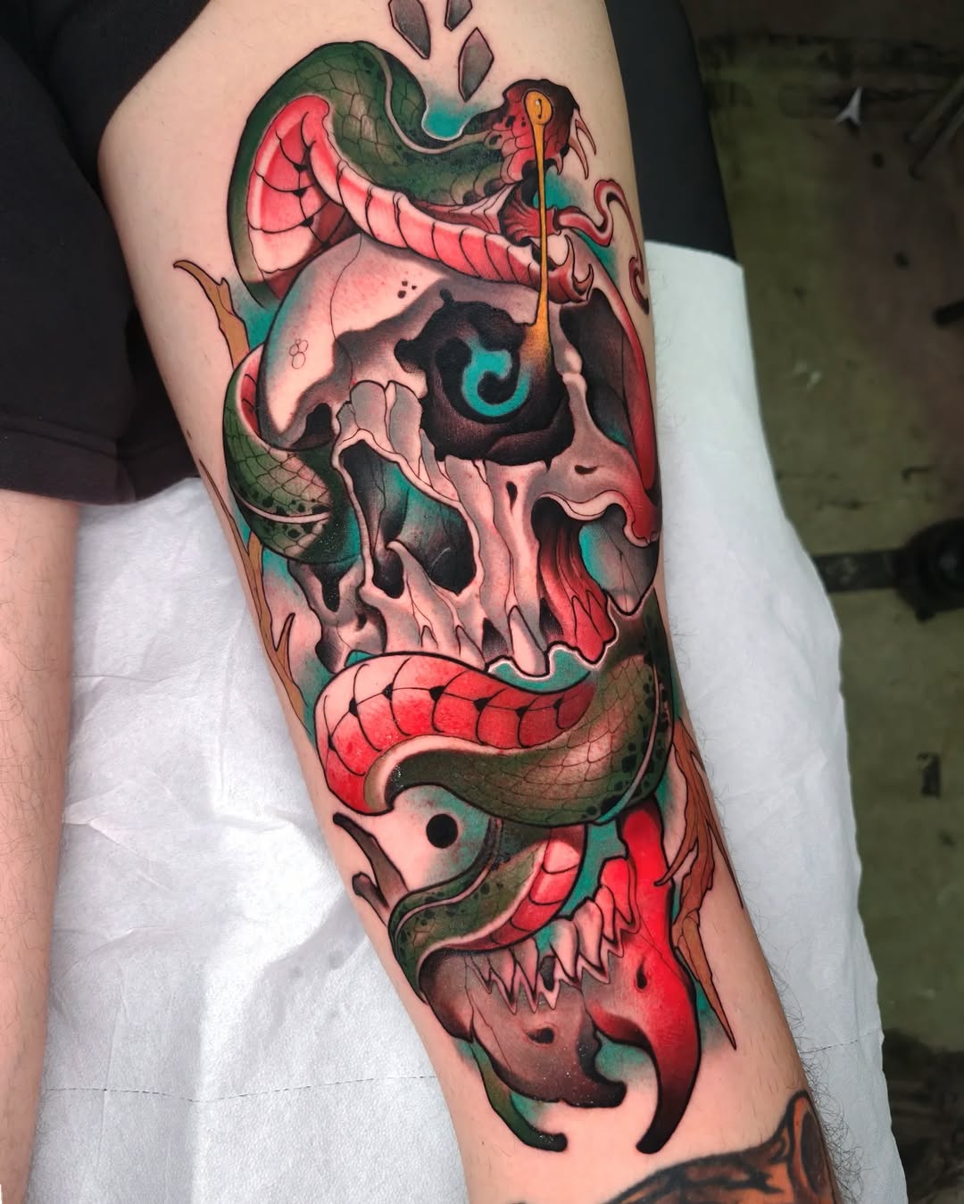Colorful skull tattoo with intertwined green snakes, red wings, and a staff with turquoise accents on the upper arm.