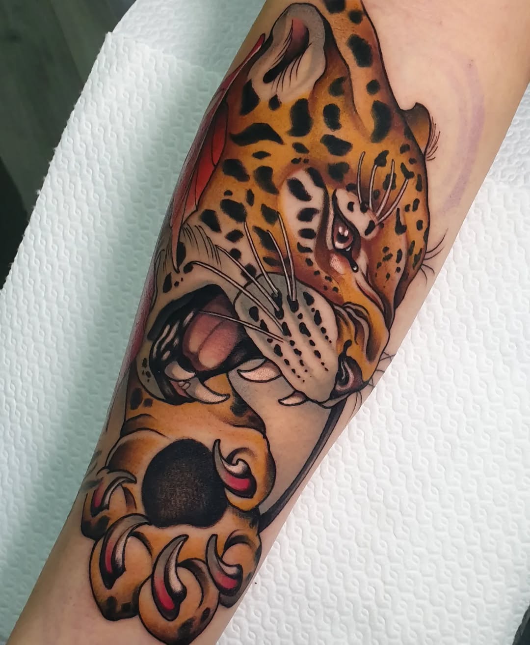 Leopard head tattoo on forearm featuring orange and black spotted fur, detailed facial features, and decorative paw designs at the wrist in red and gold tones.