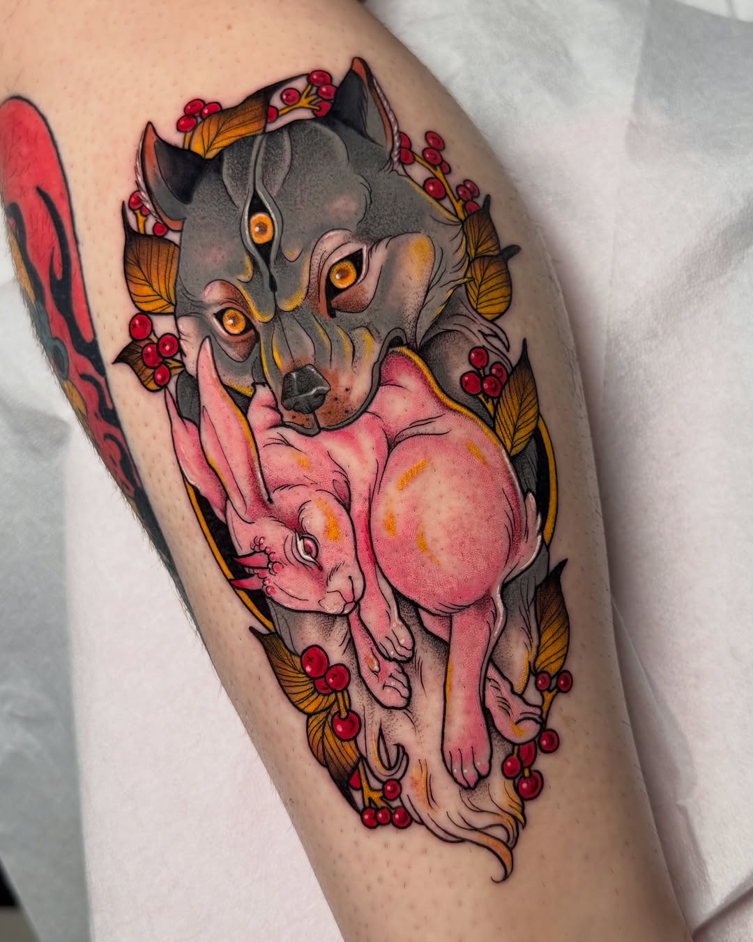Colorful tattoo on a thigh featuring a gray cat with golden eyes holding a pink pig, surrounded by red berries and golden leaves.