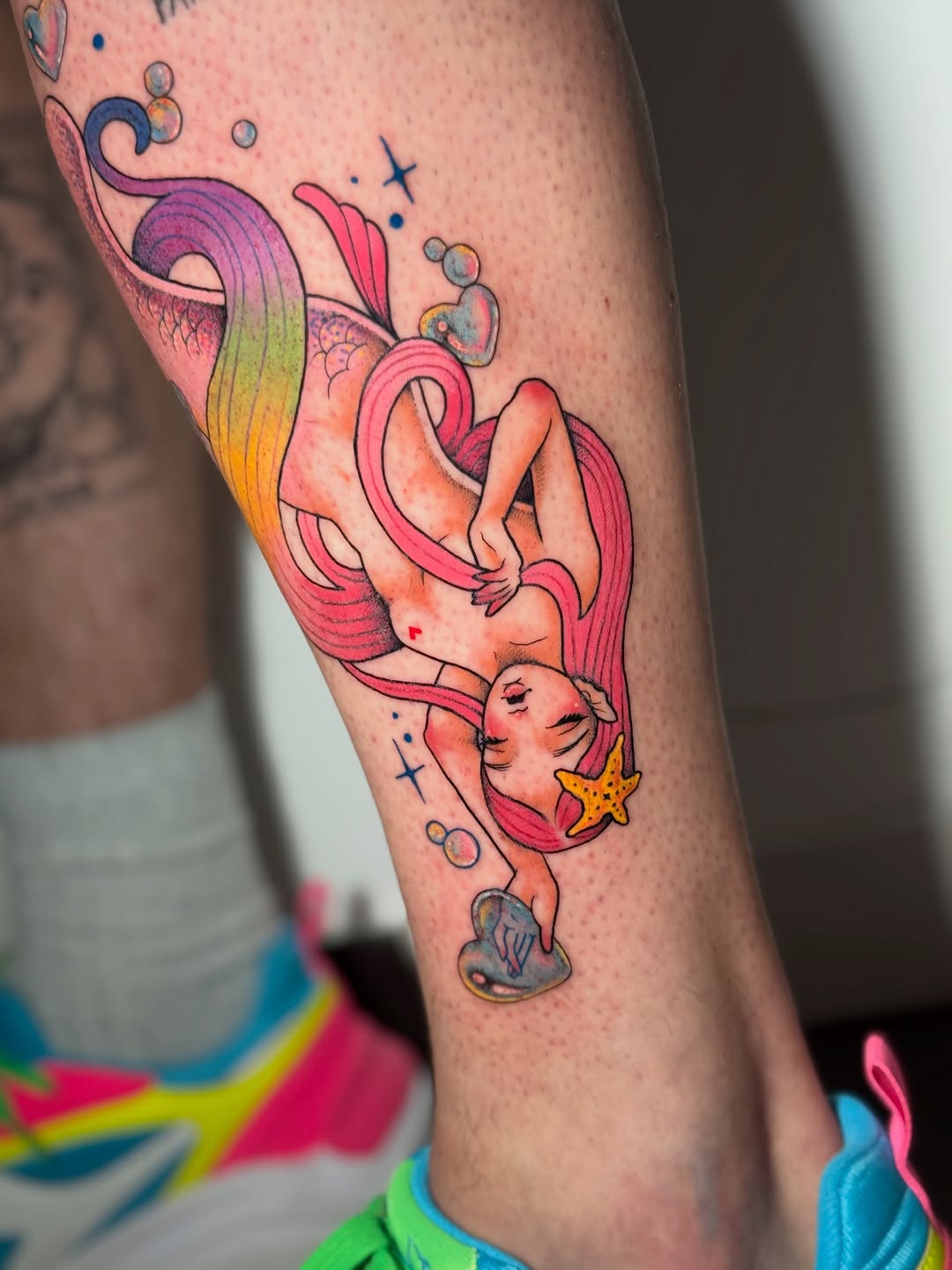 Mermaid with flowing pink and orange hair holding a starfish, surrounded by bubbles and small stars, tattooed on the calf.