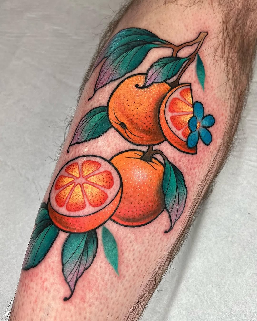 Colorful oranges and citrus slices with teal leaves and blue flowers tattooed on the forearm, featuring orange, red, and green hues.