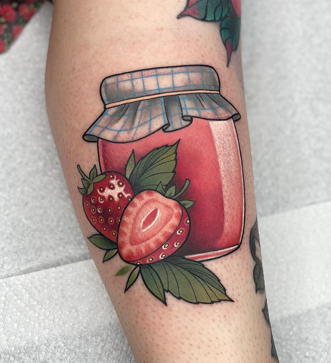 Strawberry jam jar tattoo on an arm with red jar, checkered cloth lid, fresh strawberries with green leaves, and black outline details.