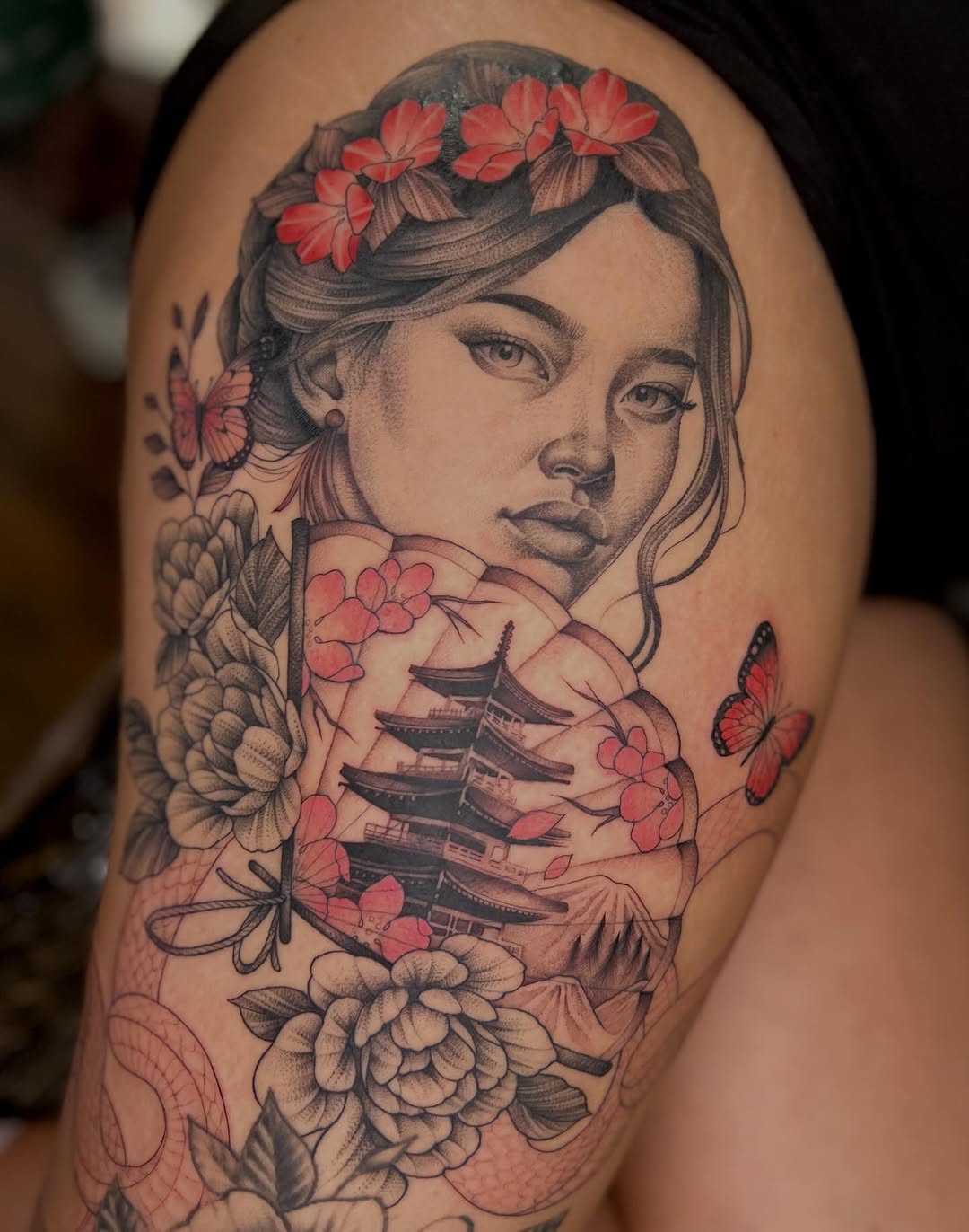 Portrait tattoo of a young woman with flower crown and pagoda imagery on her chest and torso, featuring red flowers, butterflies, and botanical elements on tan skin.