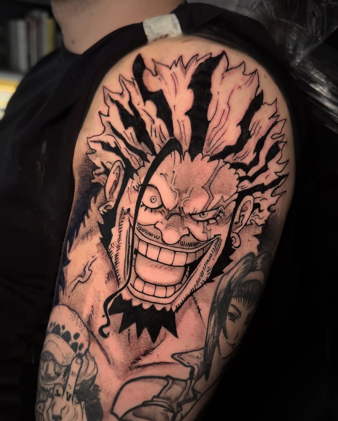 Grinning demon head tattoo with spiky hair and open mouth on upper arm, rendered in black ink with shading on tan skin.