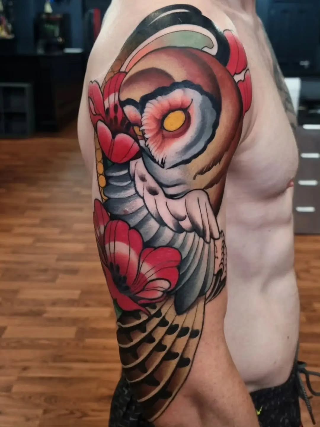 Colorful owl tattoo on upper leg featuring red flowers, yellow eyes, gray and black feathering, displayed on pale skin.