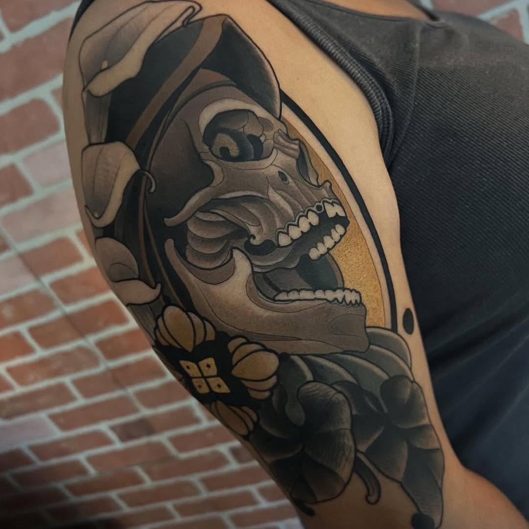Skull with open jaw wearing a wide-brimmed hat, surrounded by floral elements in black and gray ink on the upper arm.