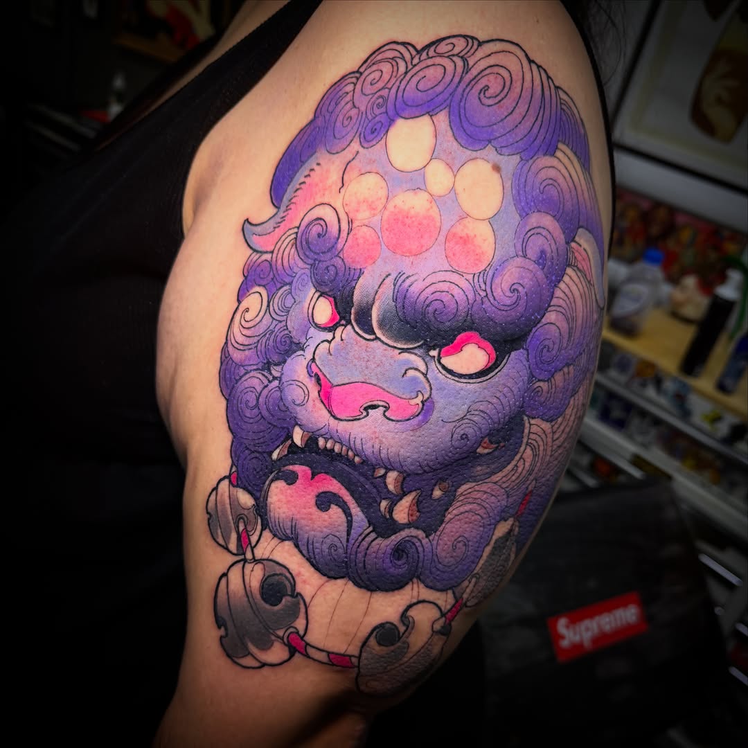 Colorful shoulder tattoo featuring a purple ornate deity or mask face with pink accents, circular patterns above, and decorative cloud-like elements below.
