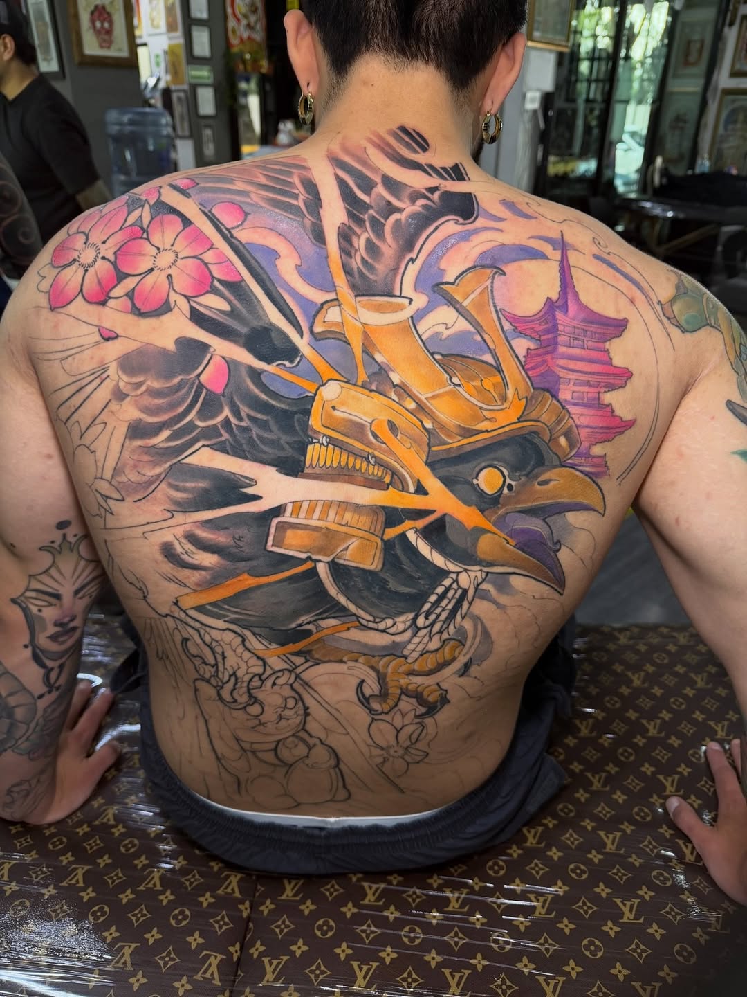 Back tattoo featuring a warrior in gold and orange armor with wings, pink flowers, purple pagoda, and decorative elements in vibrant colors.