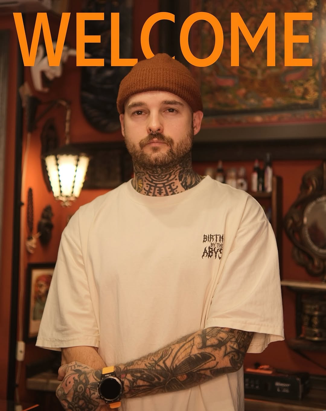 Tattooed man wearing cream t-shirt displays extensive neck and arm tattoos in black ink with geometric and figurative designs.